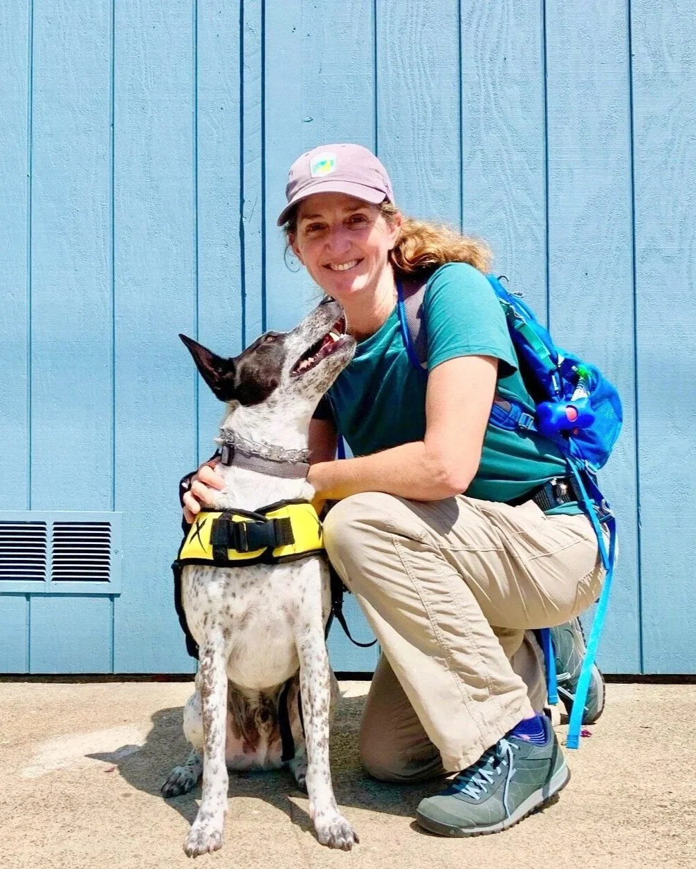 A woman in outdoor hiking gear kneels next to a black and white dog with a yellow harness, both smiling in front of a blue wooden wall.