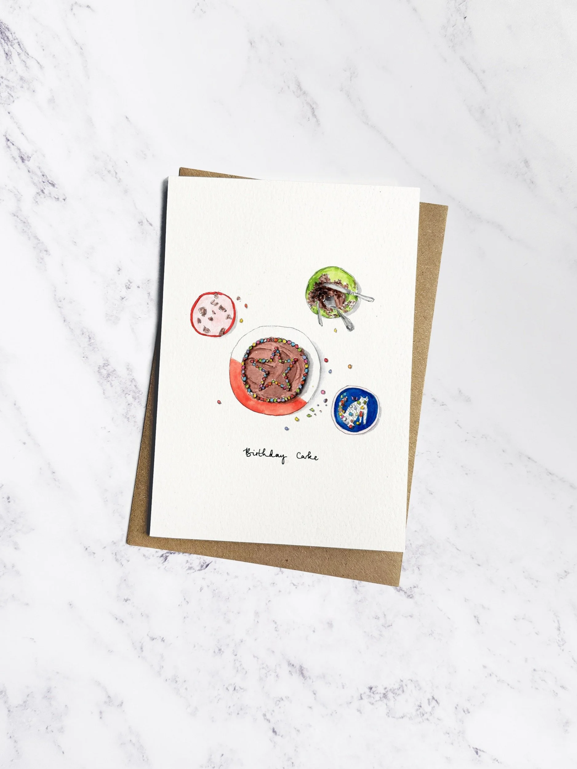 A beautifully illustrated birthday card featuring a chocolate cake with a star-shaped pattern of smarties, alongside baking tools. Displayed with a kraft envelope on a white marble background.