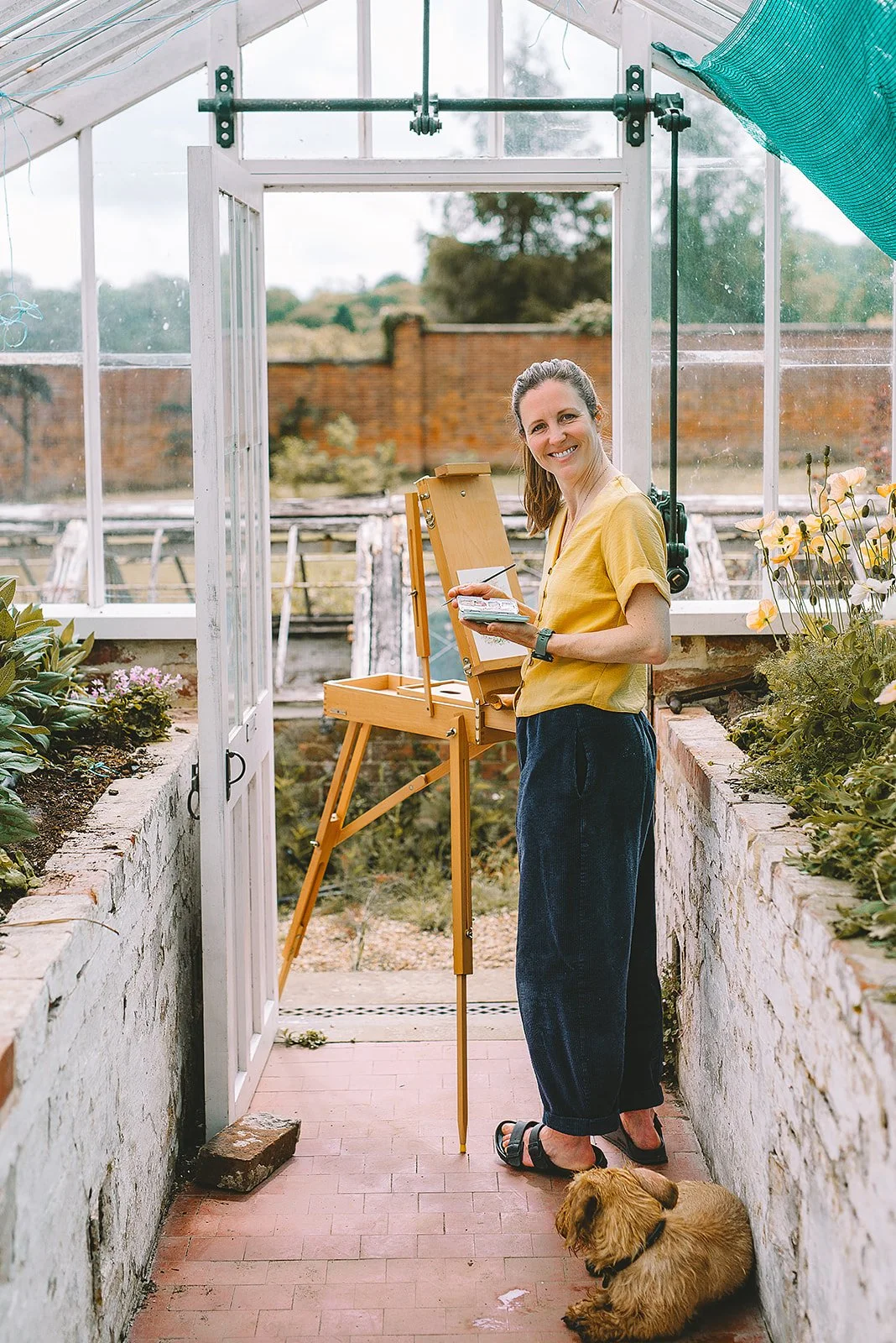 Imogen Partridge watercolour illustrator standing in a glasshouse painting at her easel standing up looking and smiling