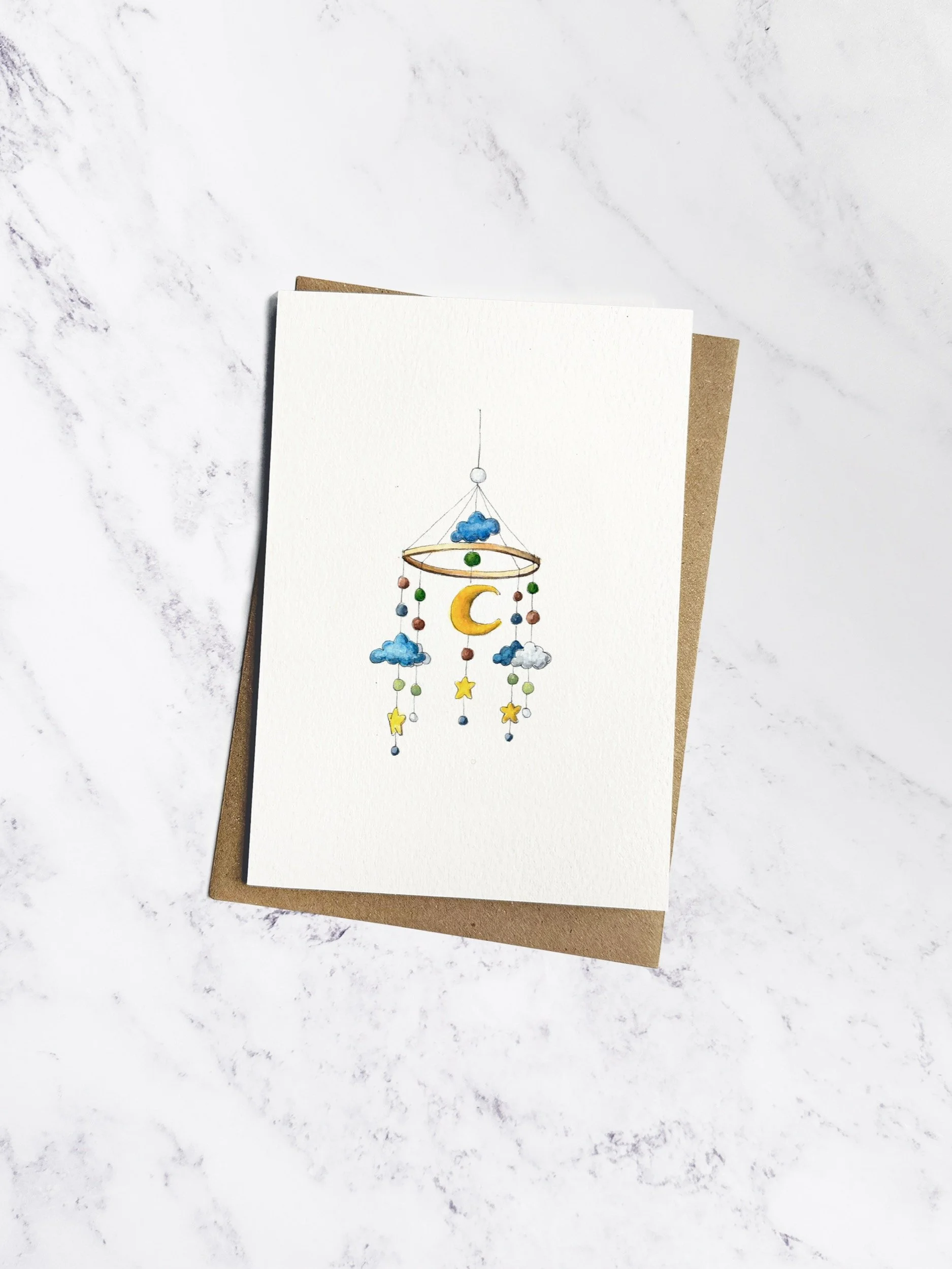 A delicate watercolour illustrated greetings card of a baby mobile featuring a moon, little hanging stars and clouds. The greetings card has a brown kraft envelope behind.