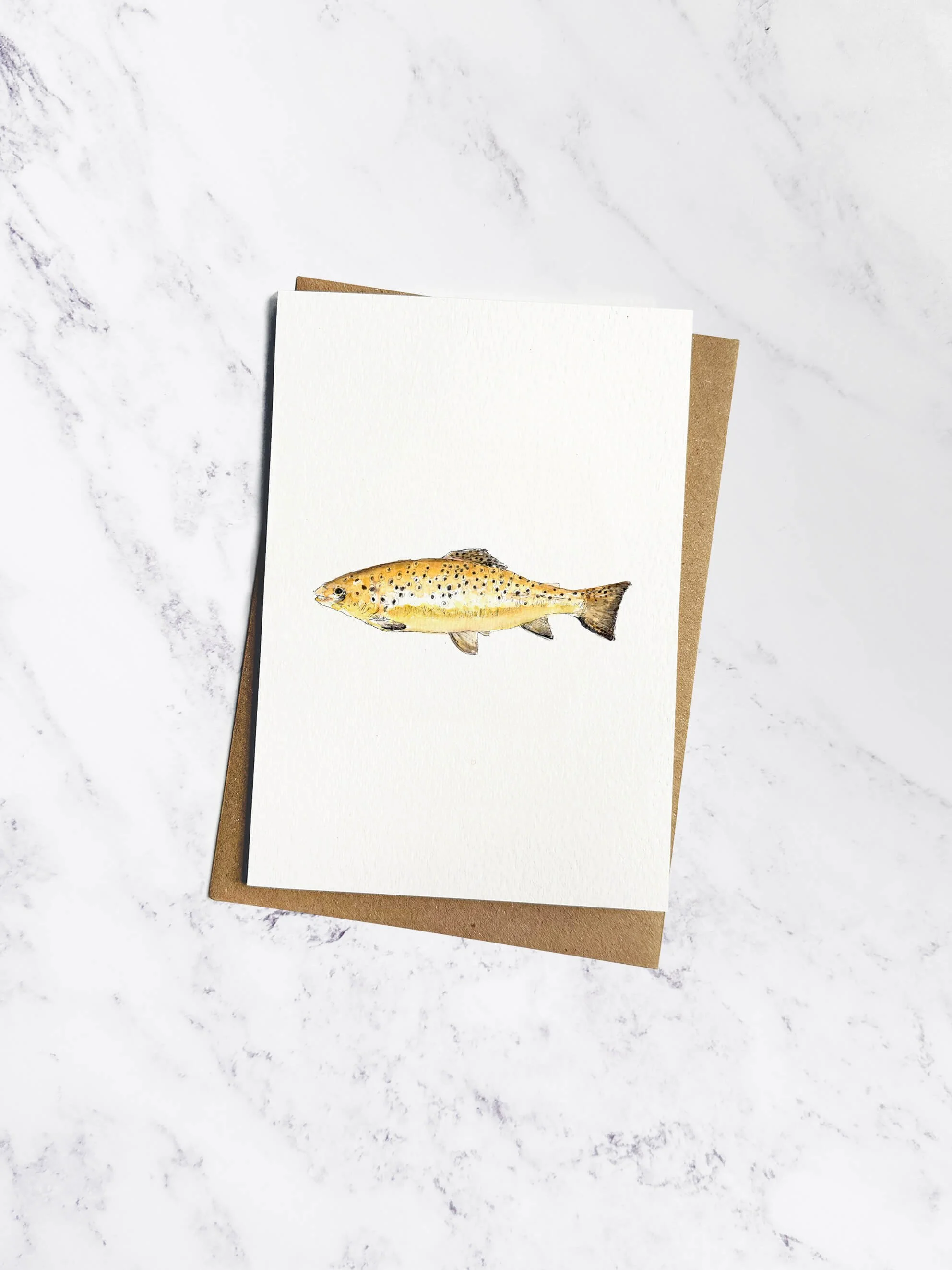A greetings card on a white marble background with beautiful watercolor illustration of a brown trout with golden and speckled patterns and a brown kraft envelope behind.