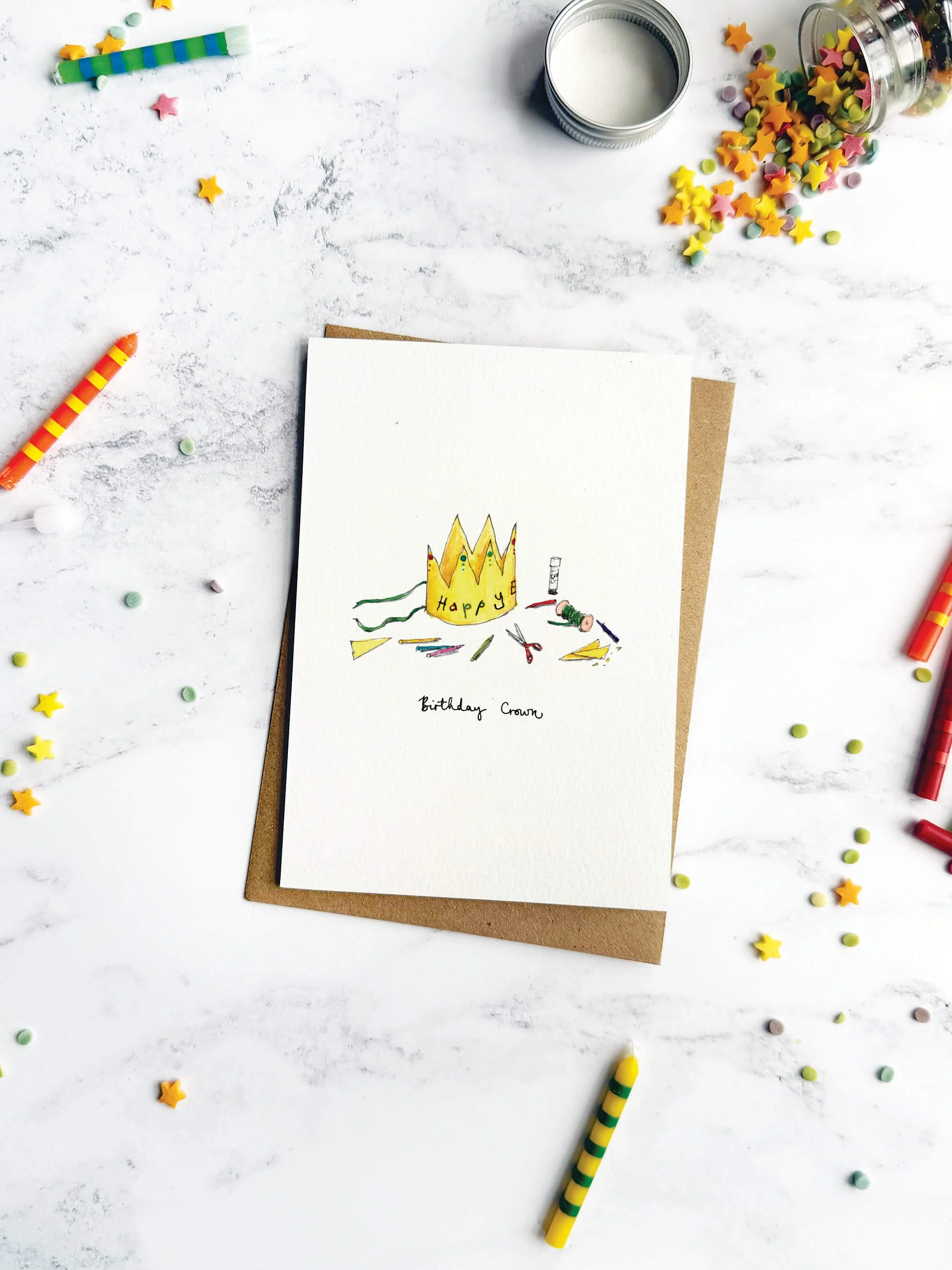 A storytelling-style birthday card featuring a DIY paper crown, styled with pastel candles and sprinkles on a marble backdrop.