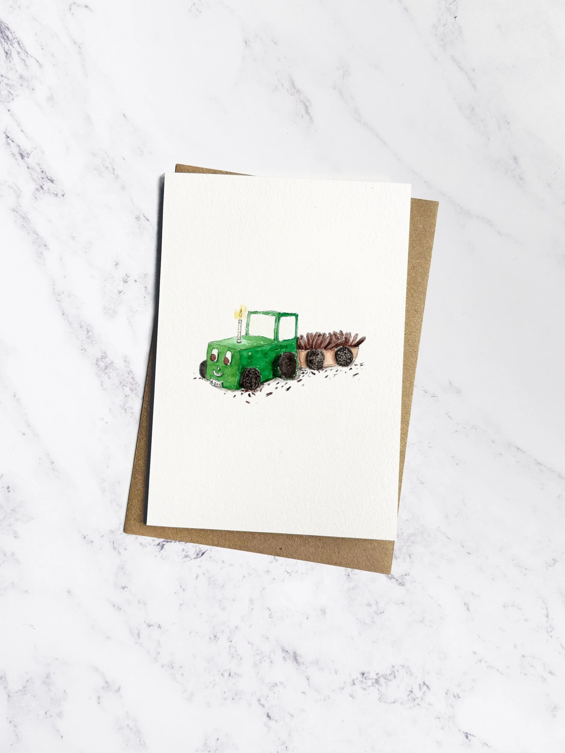 A fun illustrated birthday card featuring a homemade green tractor cake with Oreo wheels and a chocolate cupcake wheel. Displayed with a kraft envelope on a white marble background.
