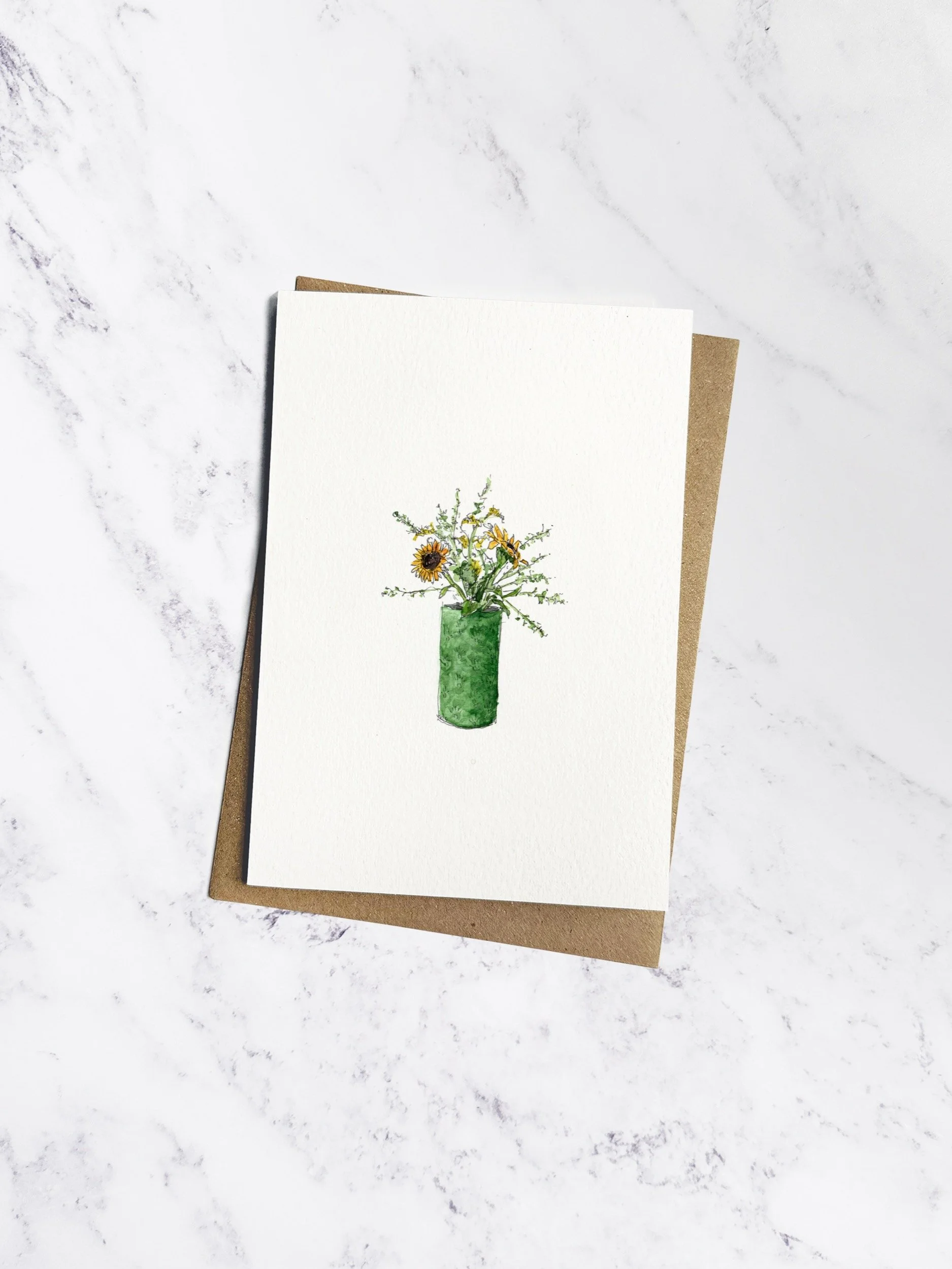 Beautifully hand painted watercolour illustration of yellow sunflowers in a green ceramic vase. The card is an off white textured paper with a brown kraft envelope behind on a marble background.