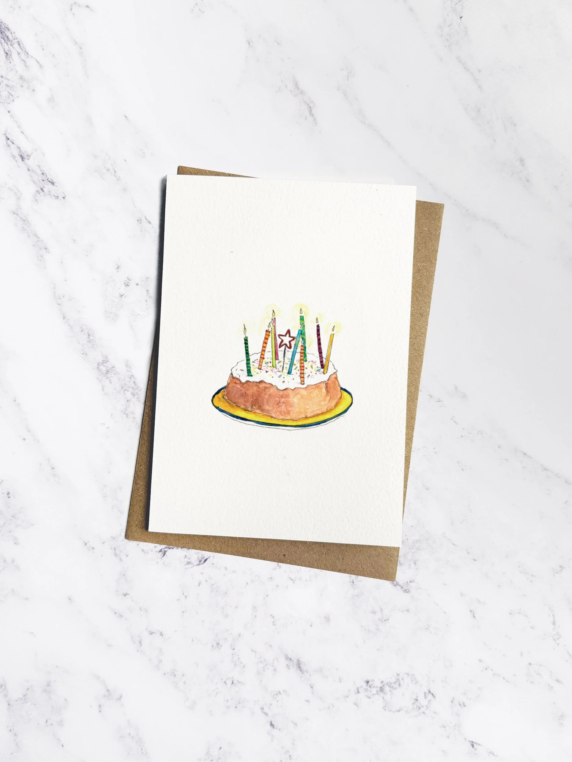 A sweet illustrated birthday card featuring a homemade sponge cake with icing, colorful sprinkles, and slightly wonky candles. Displayed with a kraft envelope on a white marble background.