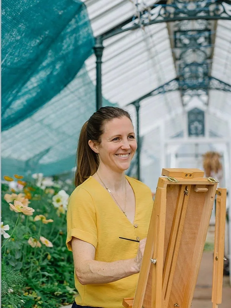 Imogen Partridge watercolour illustrator standing at her easel brush in hand smiling forward in a victorian glasshouse