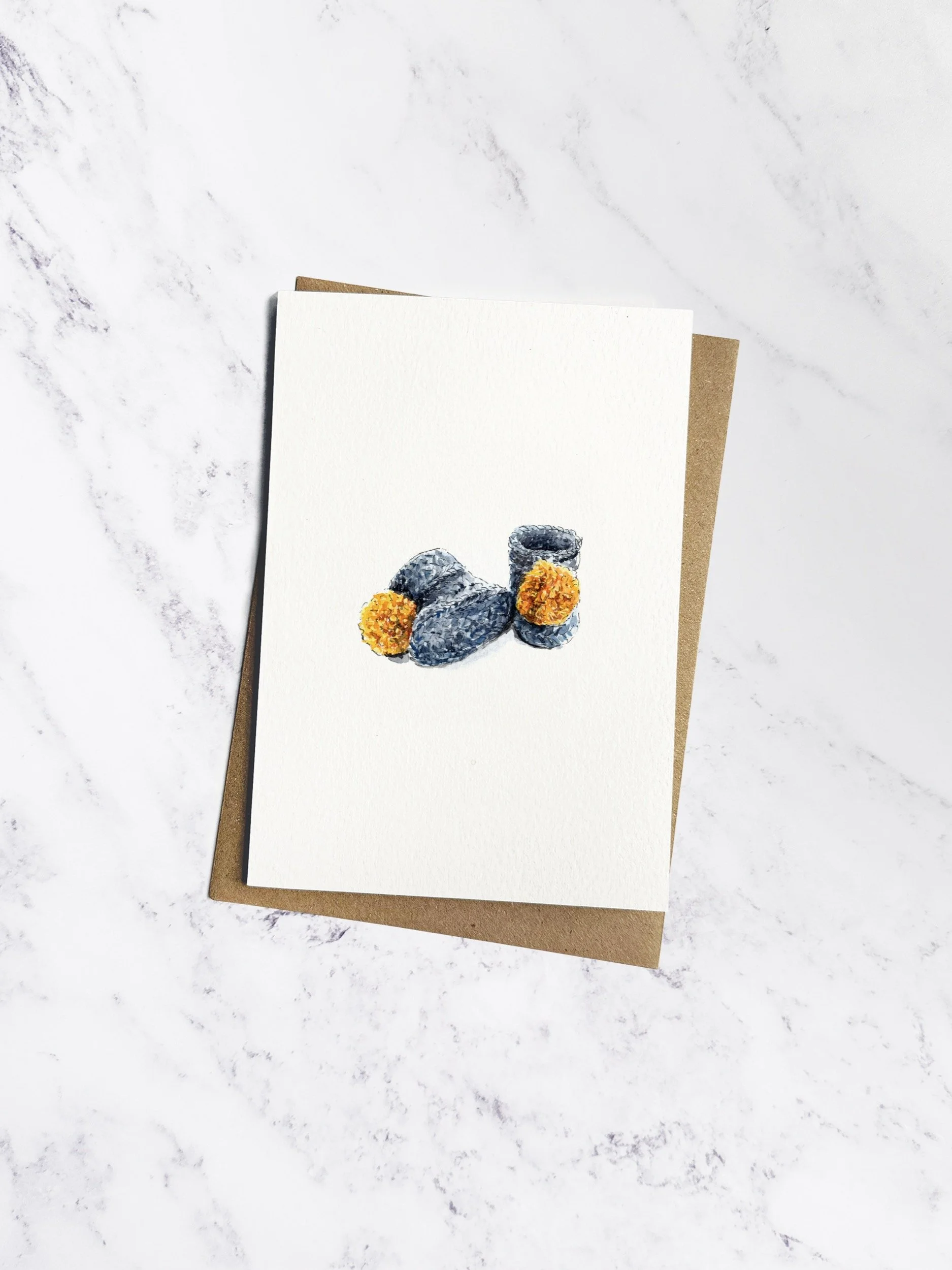 A watercolour illustrated greetings card of a pair of crochet baby booties in soft grey, with fluffy yellow pom poms on the front. One boot stands upright while the other rests on its side.