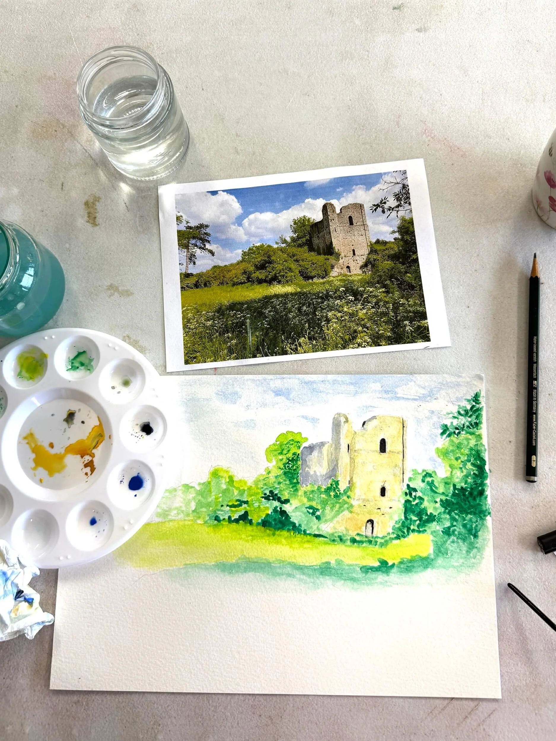 Overhead photo of a painting in progress with a reference image above showing an old tower buit in brick and green brushes and trees growing up around it with a meadow full of cow parsley in the foreground