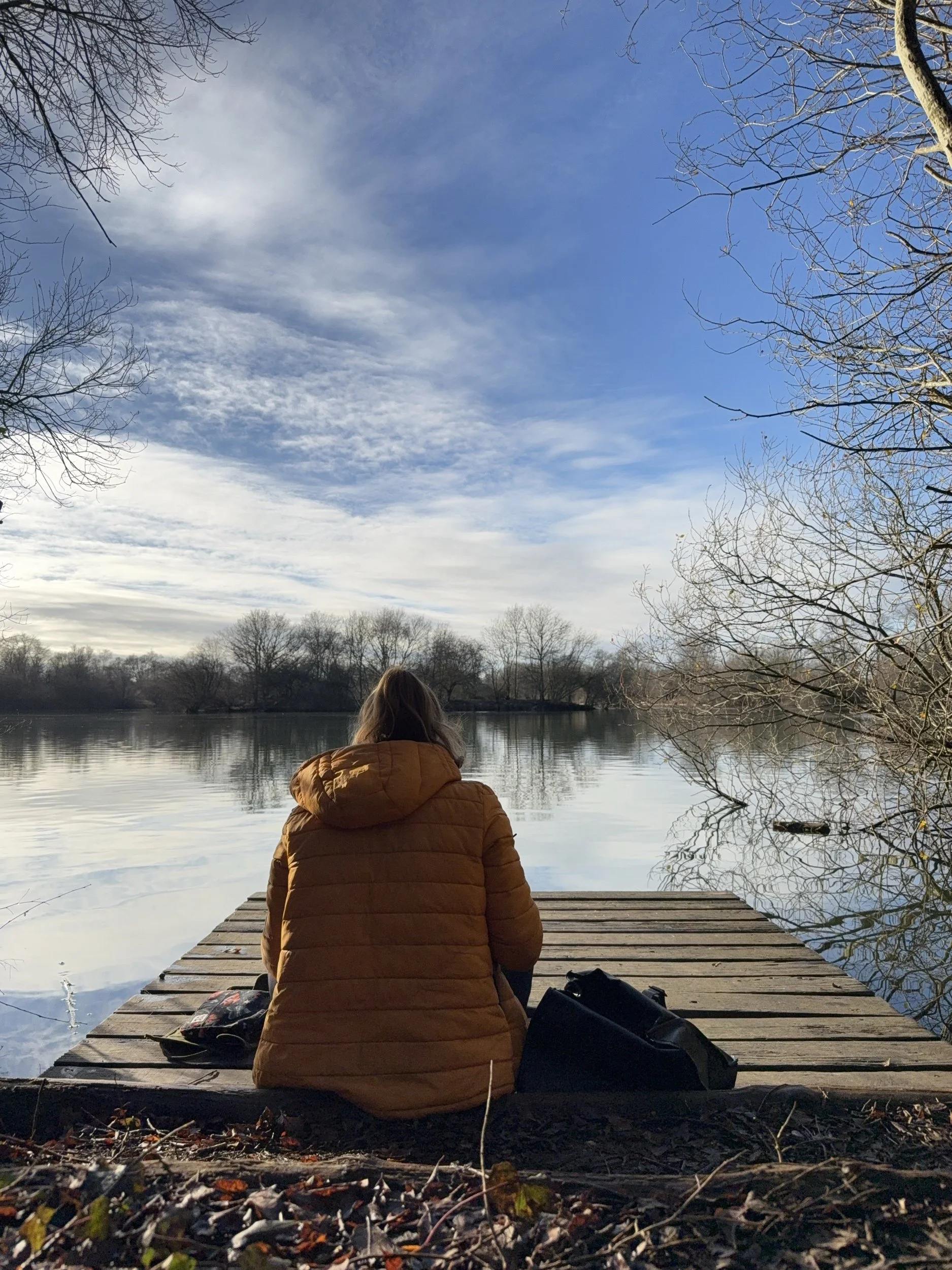 Imogen Partridge sits drawing with her back to the photo on the edge of a lake on a wooden jetty / platform. She wears a mustard yellow coat and it's winter with no leaves on trees in the distance. The lake reflects a light grey and blue sky above.