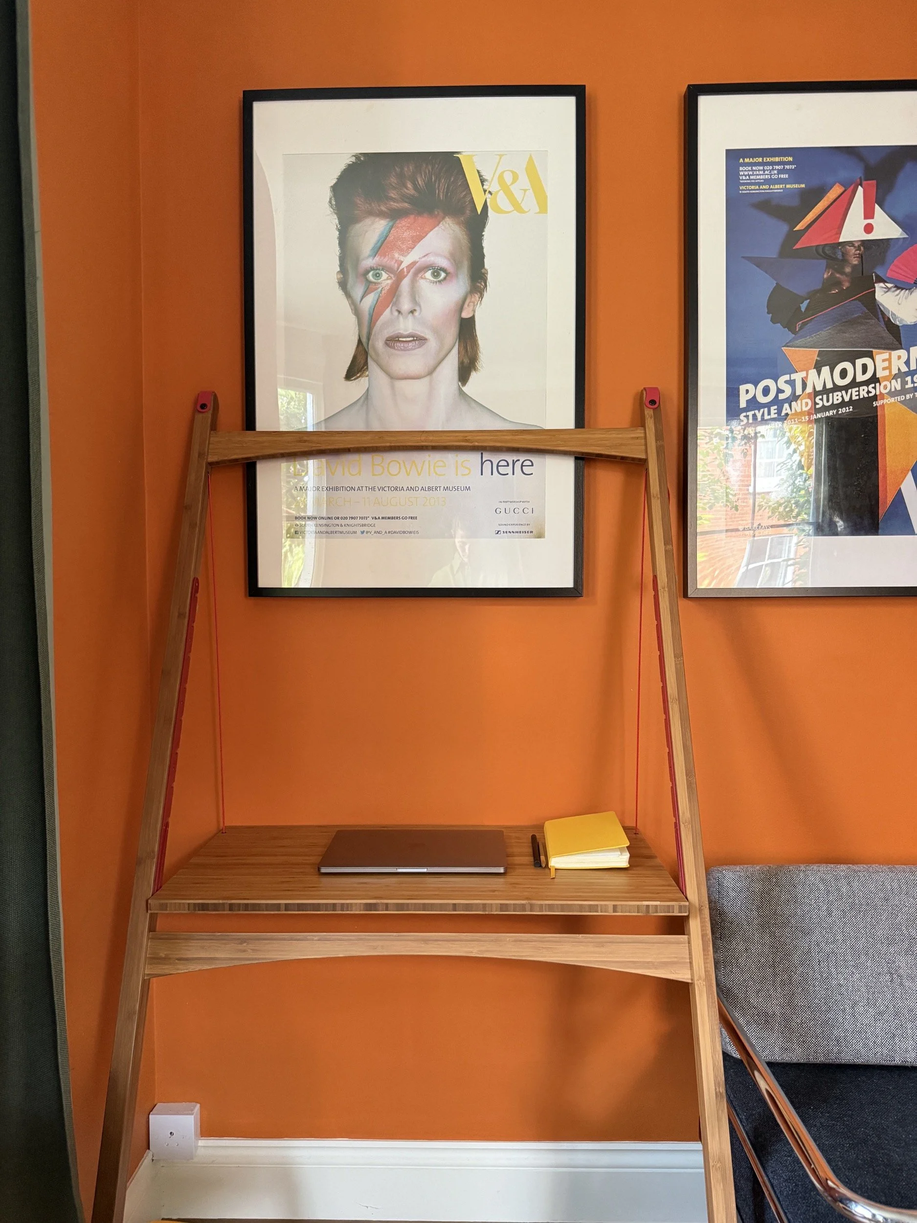 Front view of the Leandesk in an orange living from with posters on the wall. A laptop and notebook sit on the bamboo desk top