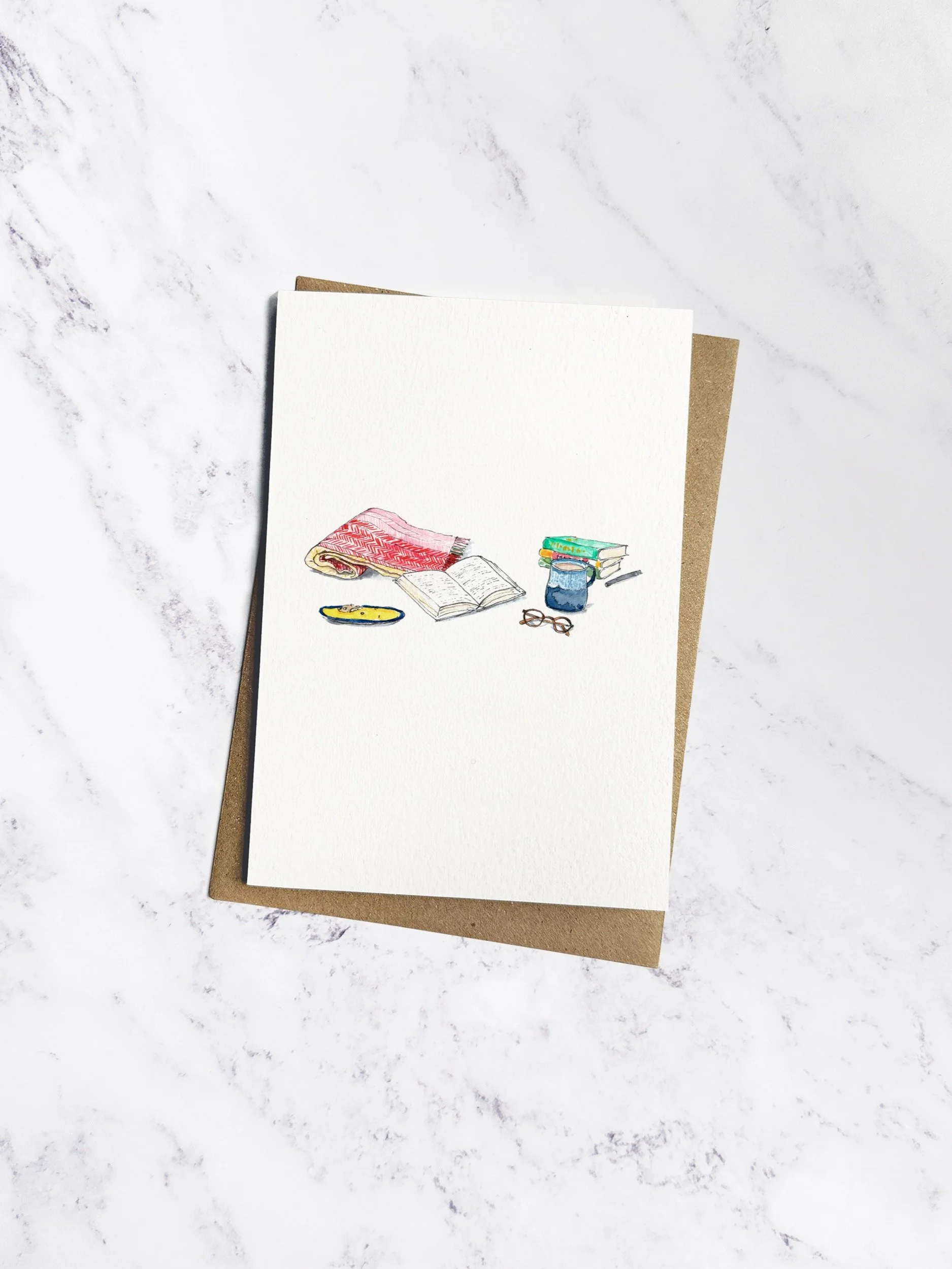 A beautifully illustrated reading-themed greetings card featuring an open book, cosy blanket, and a mug of tea. Displayed on a marble background with a kraft envelope.