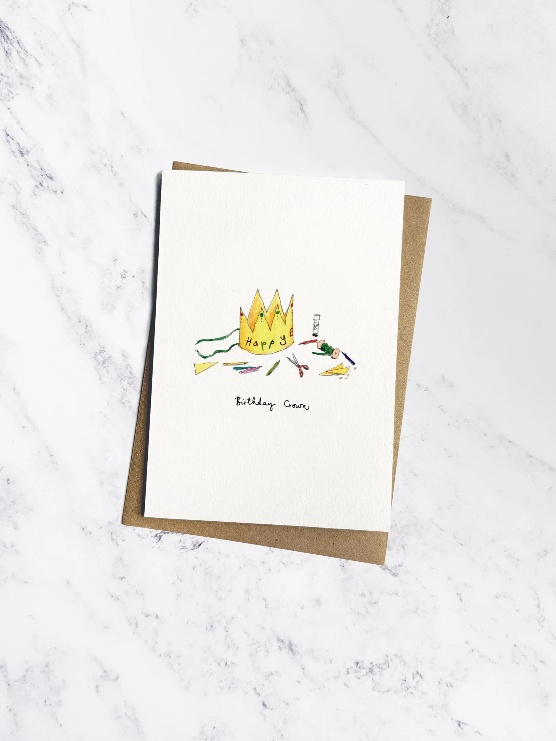 A delicate illustrated birthday card featuring a handmade yellow paper crown with ribbons and crafting materials around it. Presented with a kraft envelope on a white marble background.