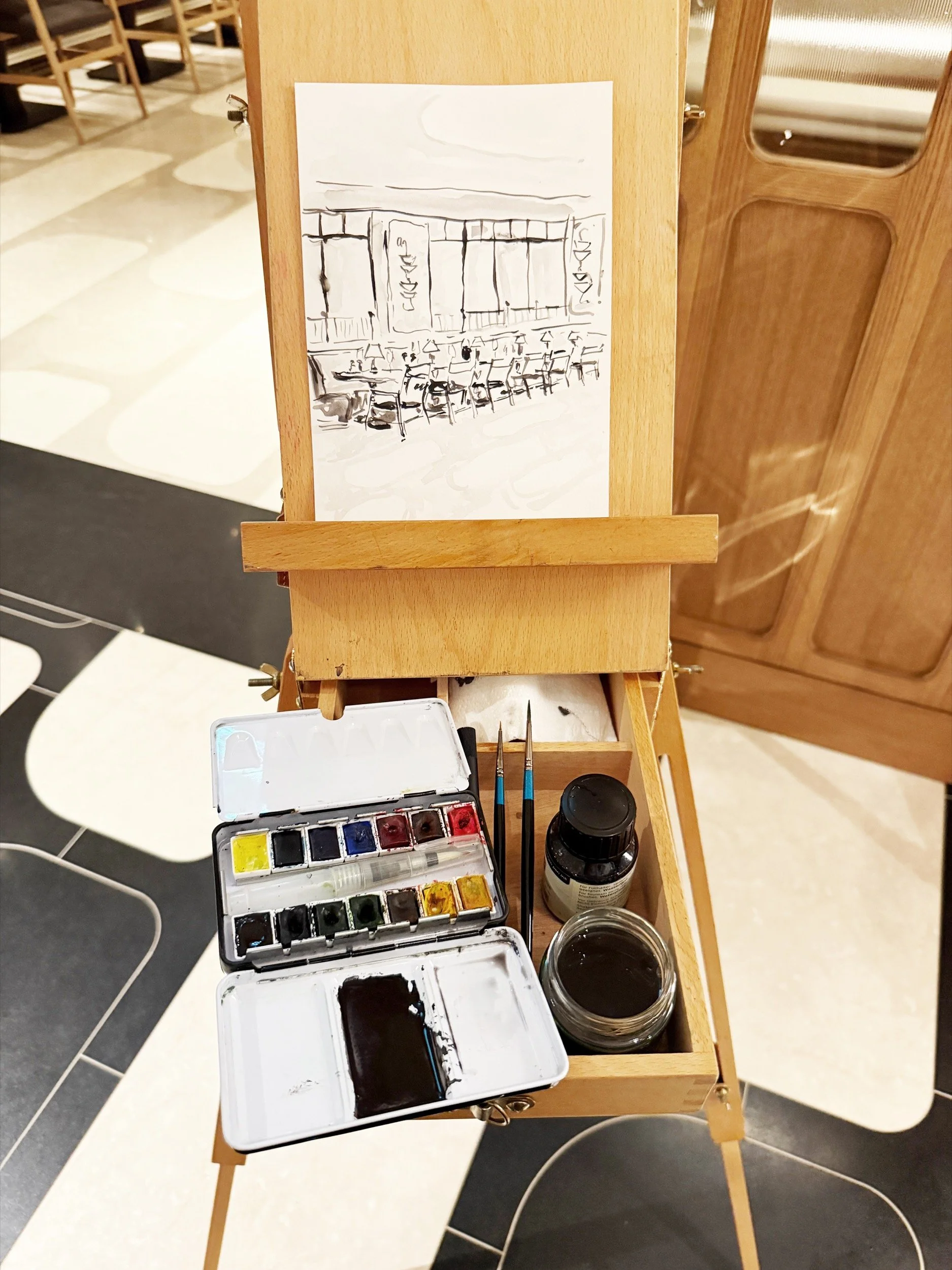 Photo showing an easel set up for live illustration at the launch of London restaurant Brasserie Angelica at The Newman in London. Monochrome line drawing and paint palette sit on the wooden easel.