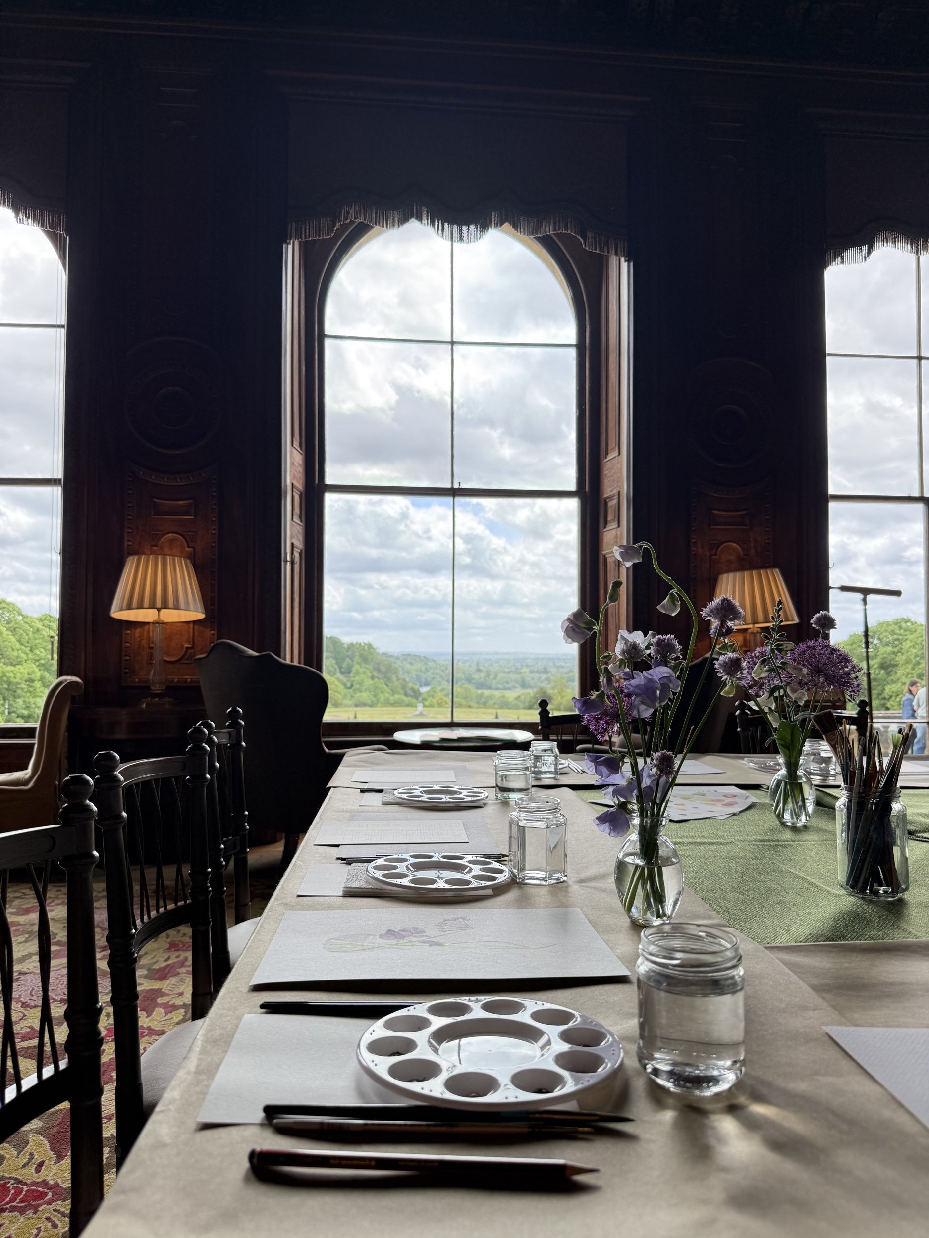 Watercolour workshop setup at Cliveden National Trust. Photo shows the table set up with worksheets, paper, watercolour brushes and materials for the workshop. The window beyond looks out onto the gardens and green space outside