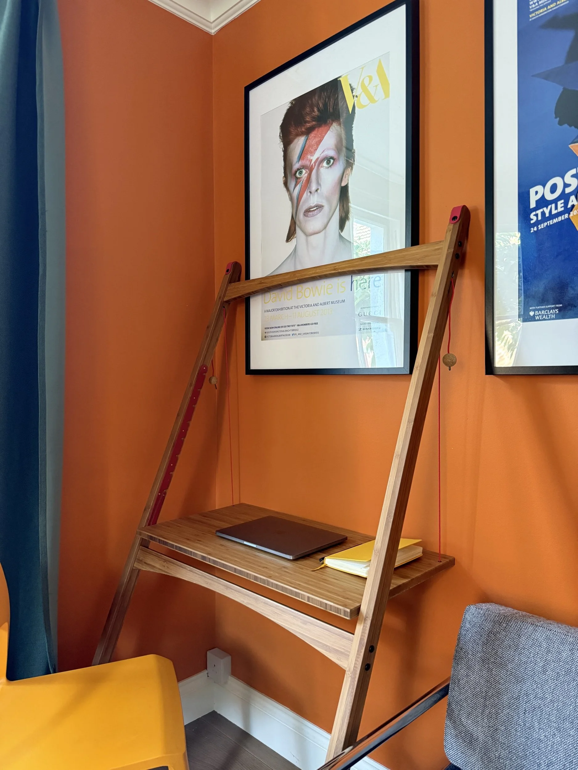 Leandesk shown in living room standing against a wall at an angle with posters framed on the wall and a yellow chair infront