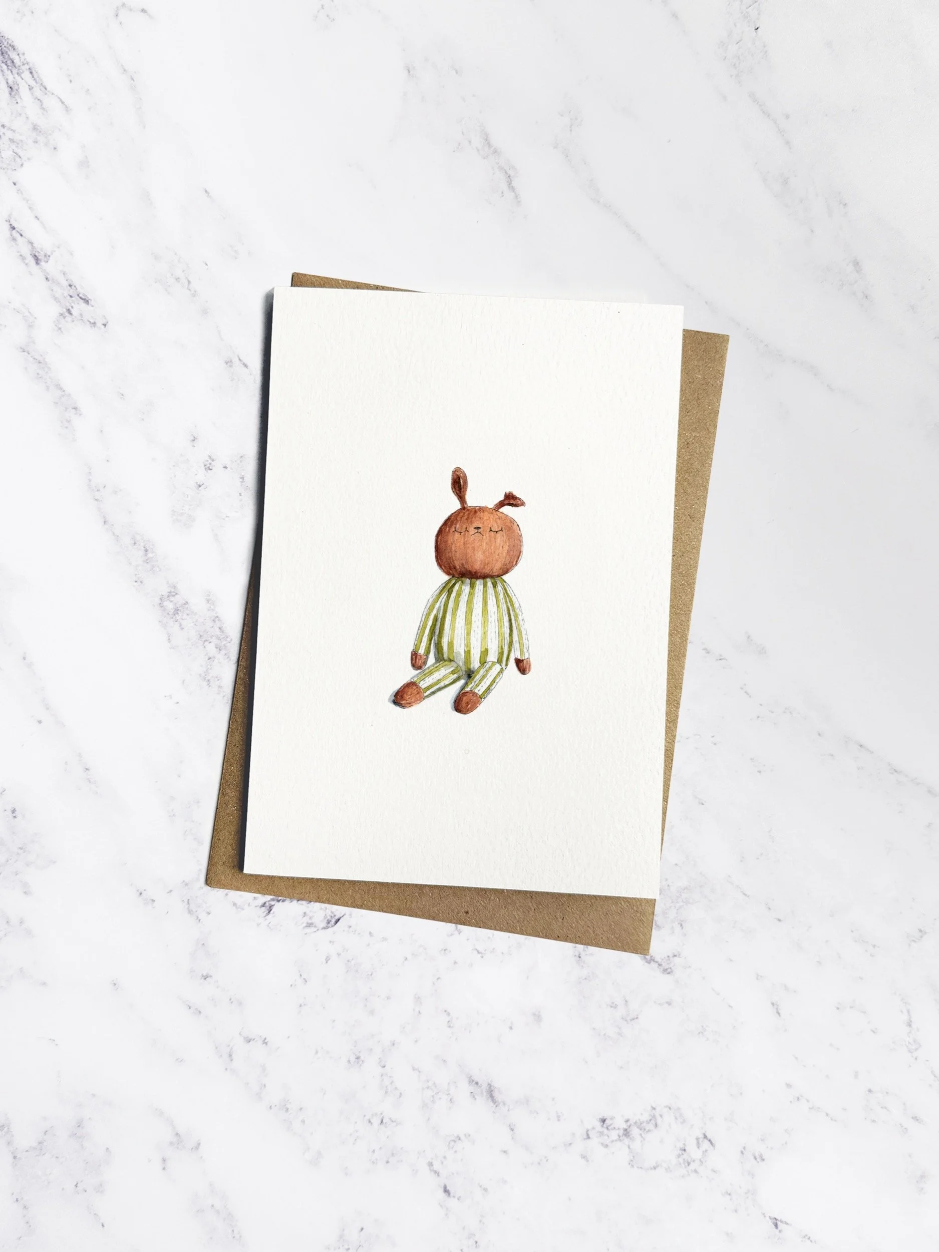 An off white greetings card, with a brown kraft envelope behind, depicting a watercolour illustration of a handmade knitted brown bunny soft toy, dressed in a stripy green and white outfit.