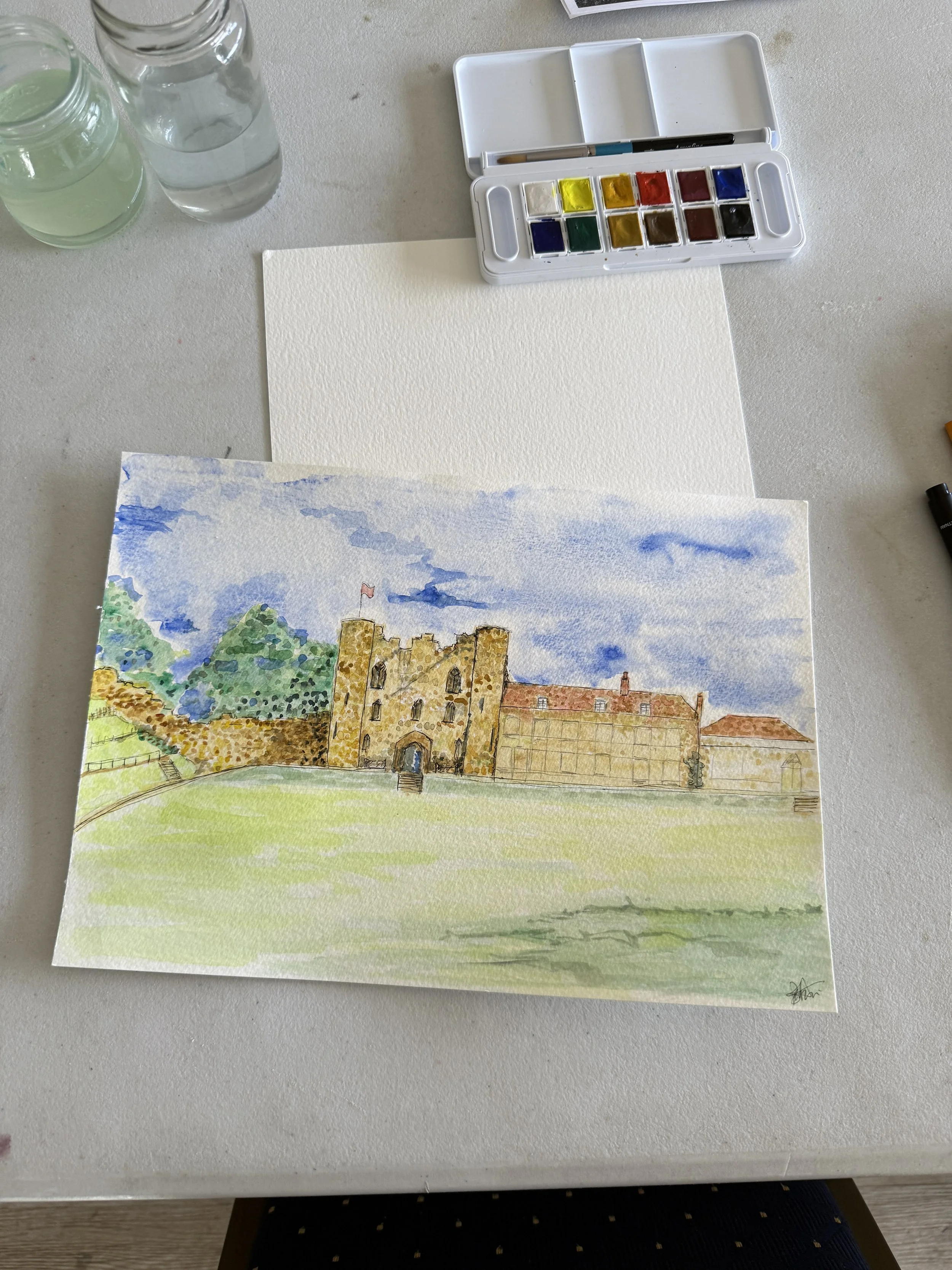 Overhead photo of a painting of Tonbridge Castle in progress. A small travel paint set sits at the top of the page with water jars to the top left. The painting is Tonbridge Castle with a green lawn at the front and blue sky in the background.