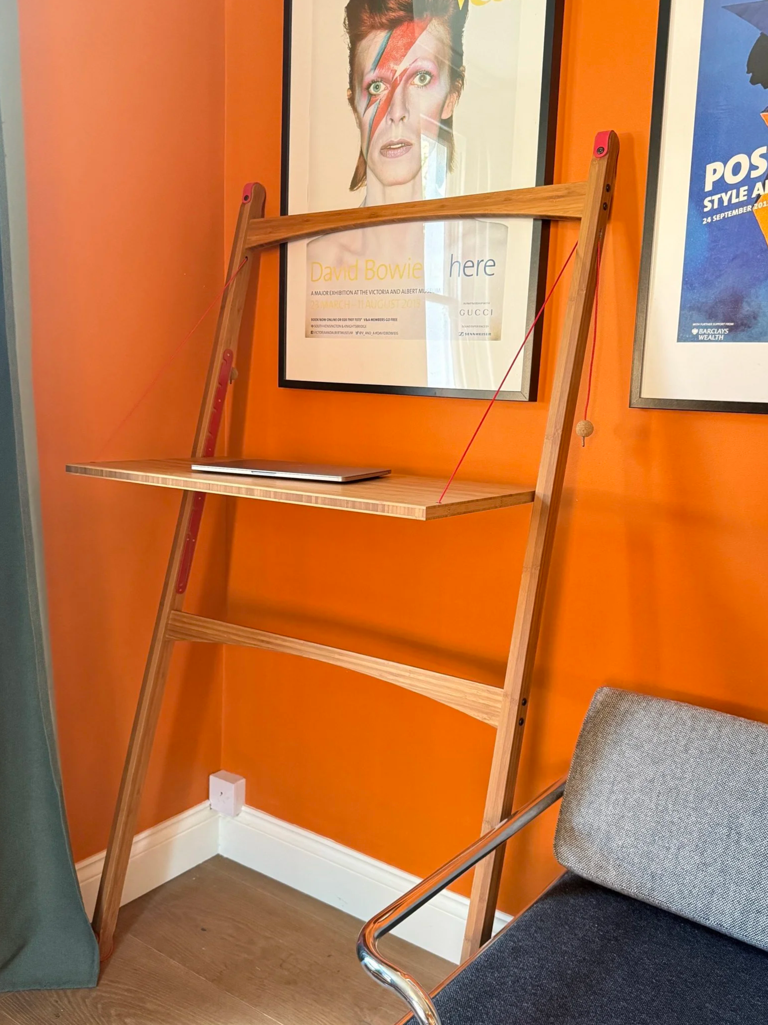 Leandesk set up against a wall in standing position. The bamboo frame leans against an orange wall with framed posters behind and a laptop sits on the desk itself