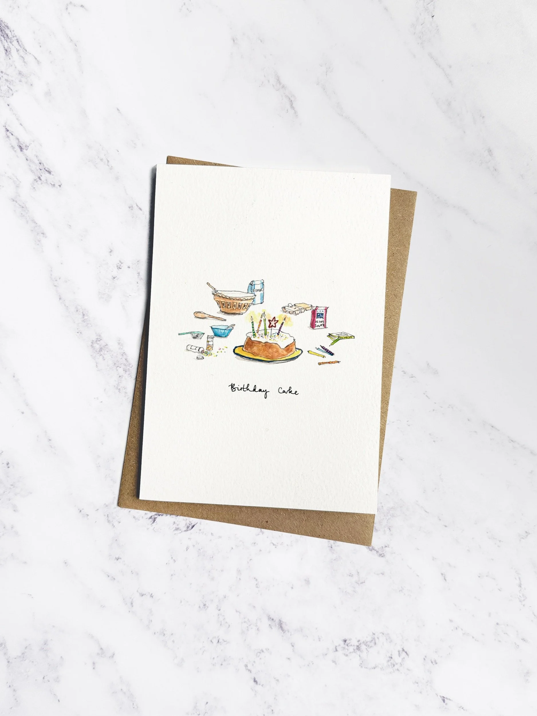 A charming illustrated birthday card featuring a homemade cake with candles, surrounded by baking. Shown with a kraft envelope on a white marble background.