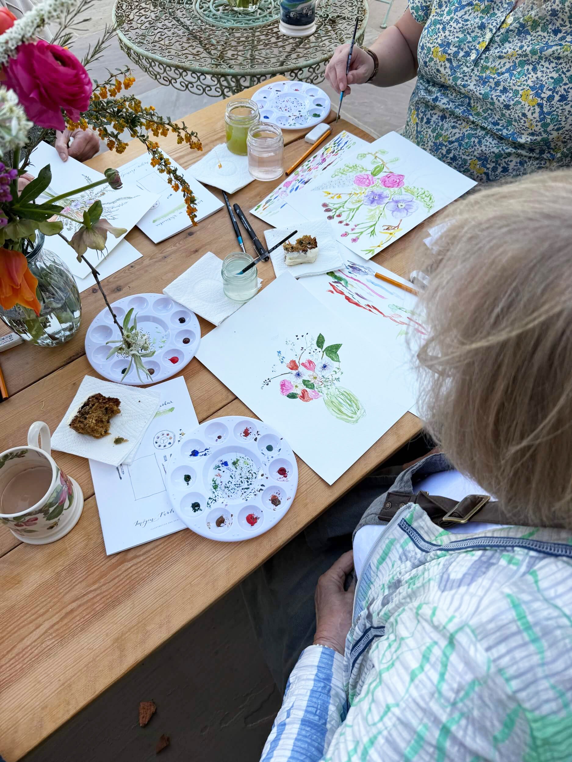 Overhead view of a wooden table covered with painting equipment and two paintings of flowers in progress. Flowers can be seen to the left of the shot and brushes, pens, paint palettes are on the table.