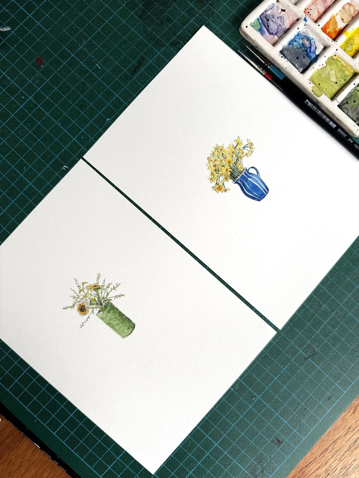 Beautifully illustrated watercolour sunflowers and daffodil designs sit on a page next to each other, with a paint palette to the right on a green cutting mat background.