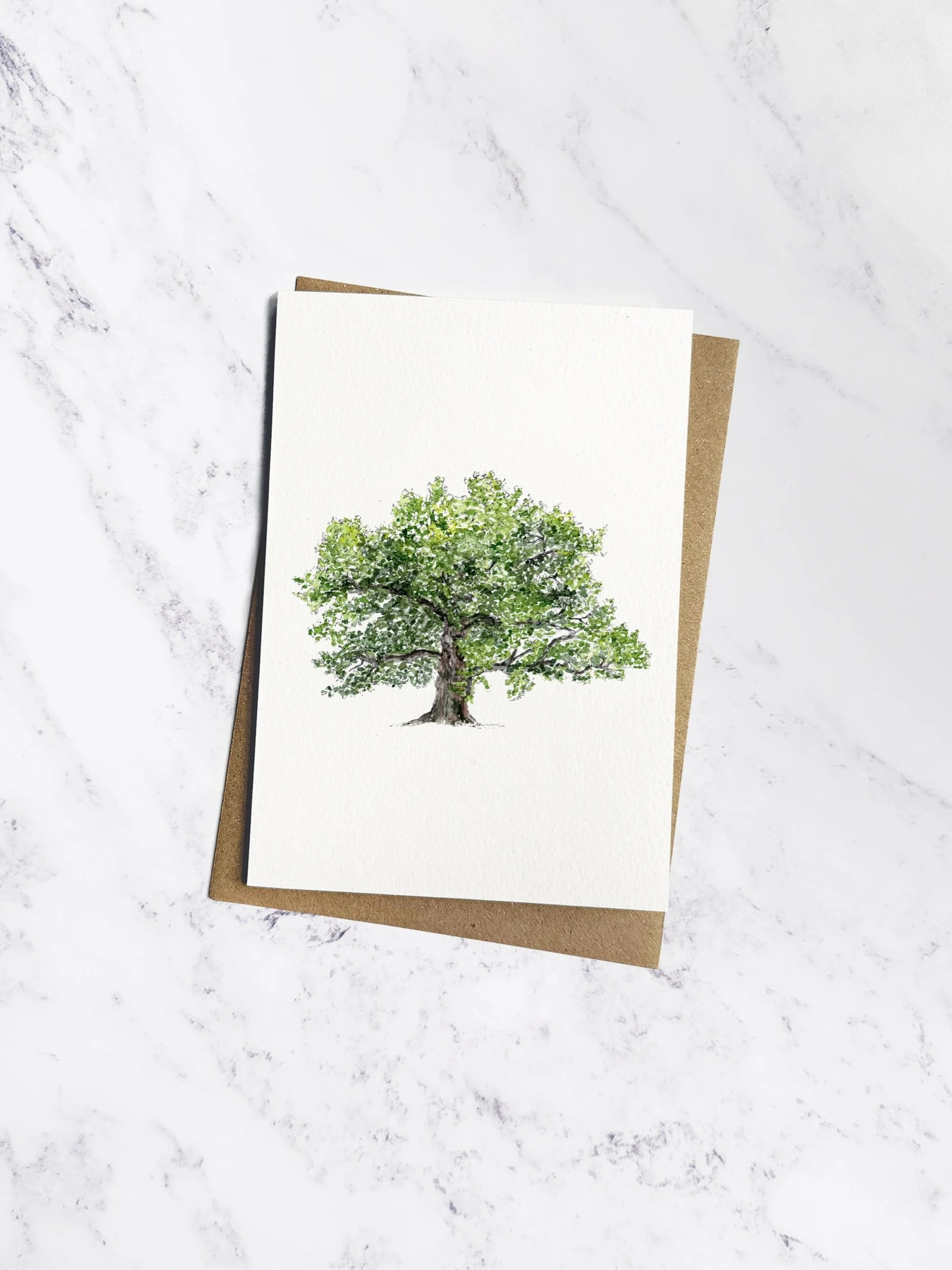 Watercolour illustration of a beautiful leafy green oak tree, on a white greeting card, a brown kraft envelope is tucked behind and it sits on a white marble background