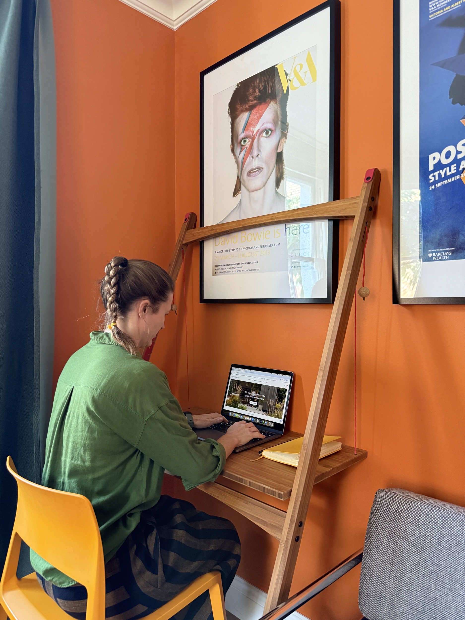 White brown haired female sitting at Leandesk on a yellow chair. Orange walls behind with bamboo desk and red cord supports.