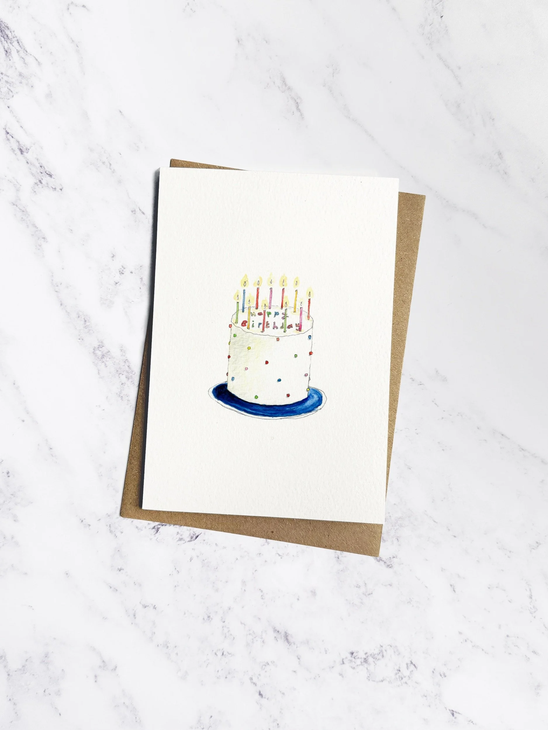 A beautifully illustrated birthday card featuring a tall, white cake with colourful icing spots, candles, & "Happy Birthday" in icing. Presented with a kraft envelope on a white marble background.