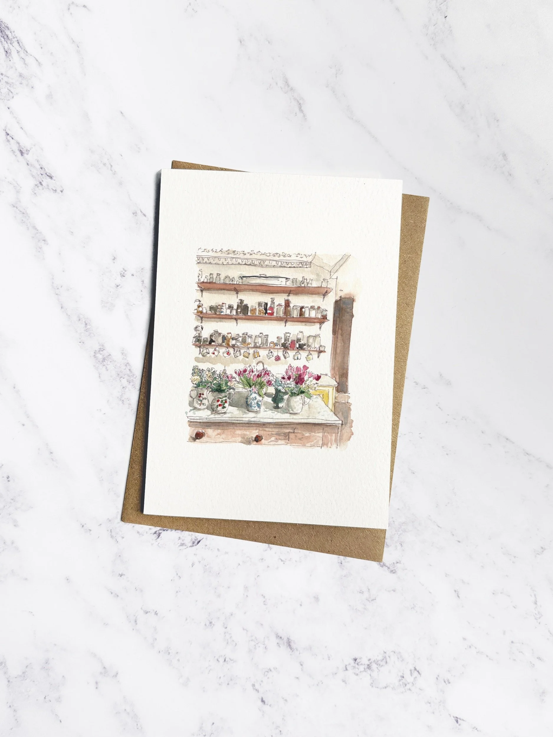 Beautifully hand painted watercolour kitchen scene with brimming jugs of tulips sitting at the front. The card is on an white textured paper with brown kraft envelope behind on a marble background.