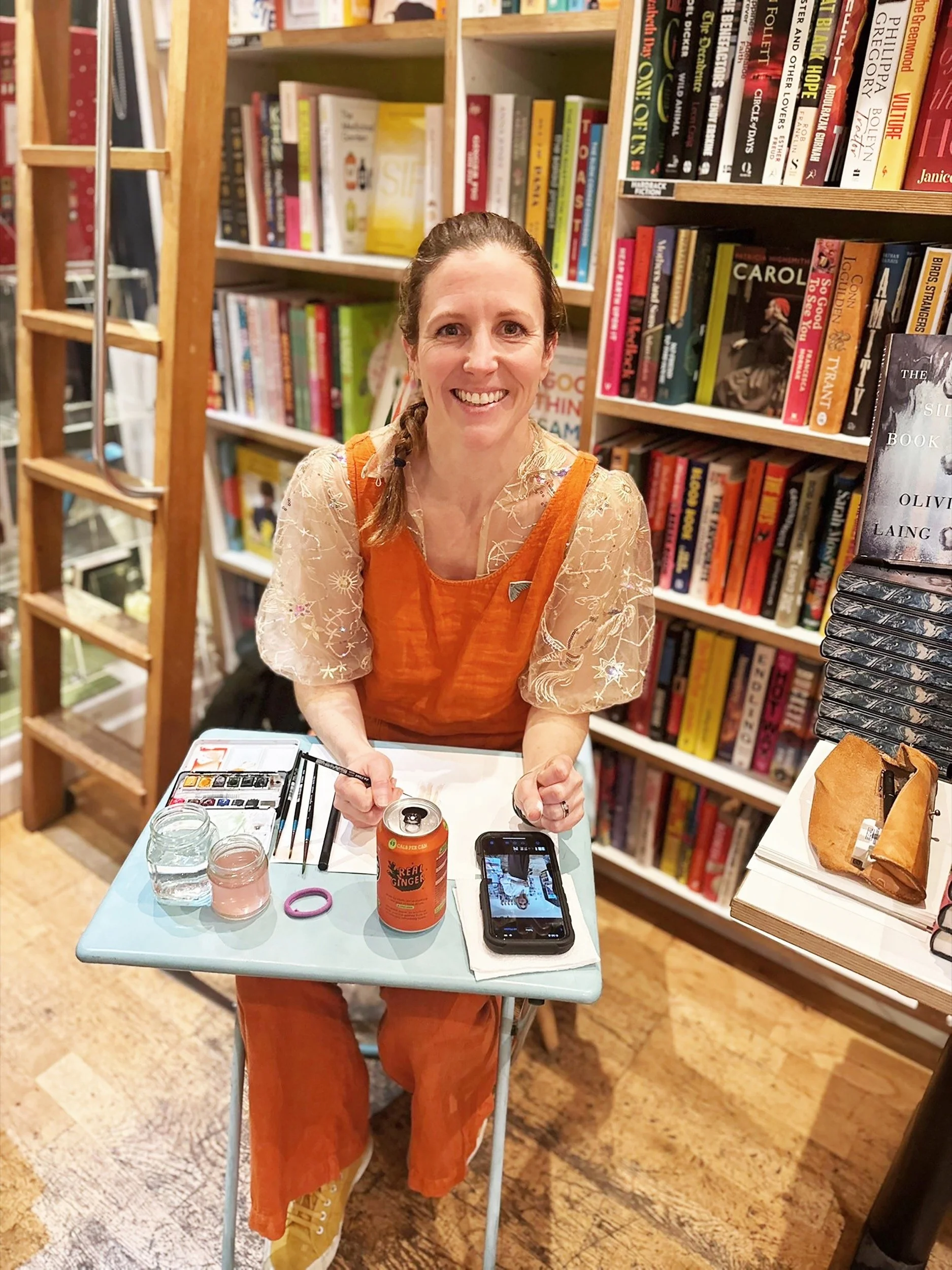 Imogen Partridge illustrating live at a book launch event in London. Sitting at a small table with watercolour paints and smiling at the camera