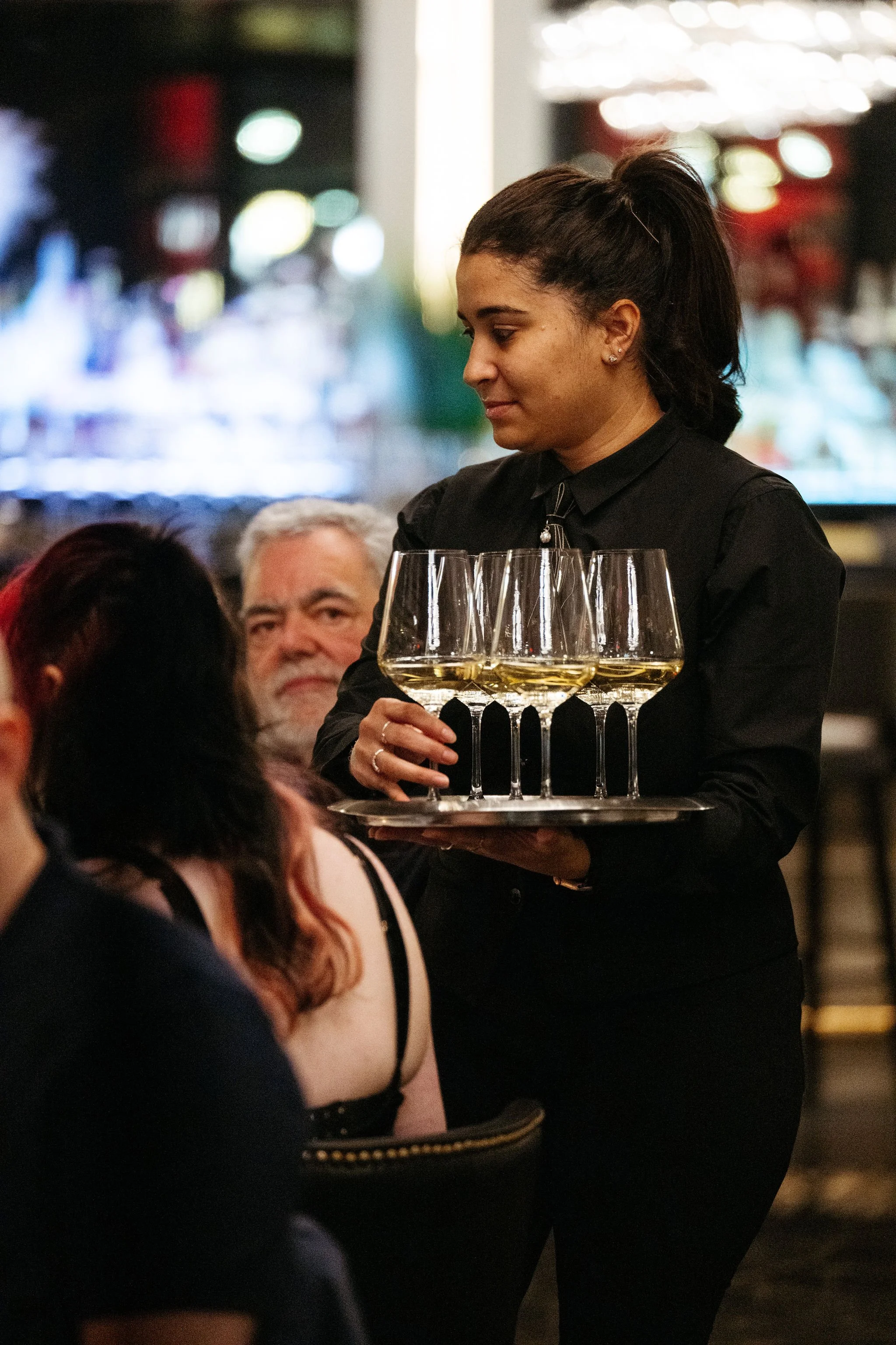 A waitress in black uniform serving a tray of four glasses of white wine at a restaurant or bar.