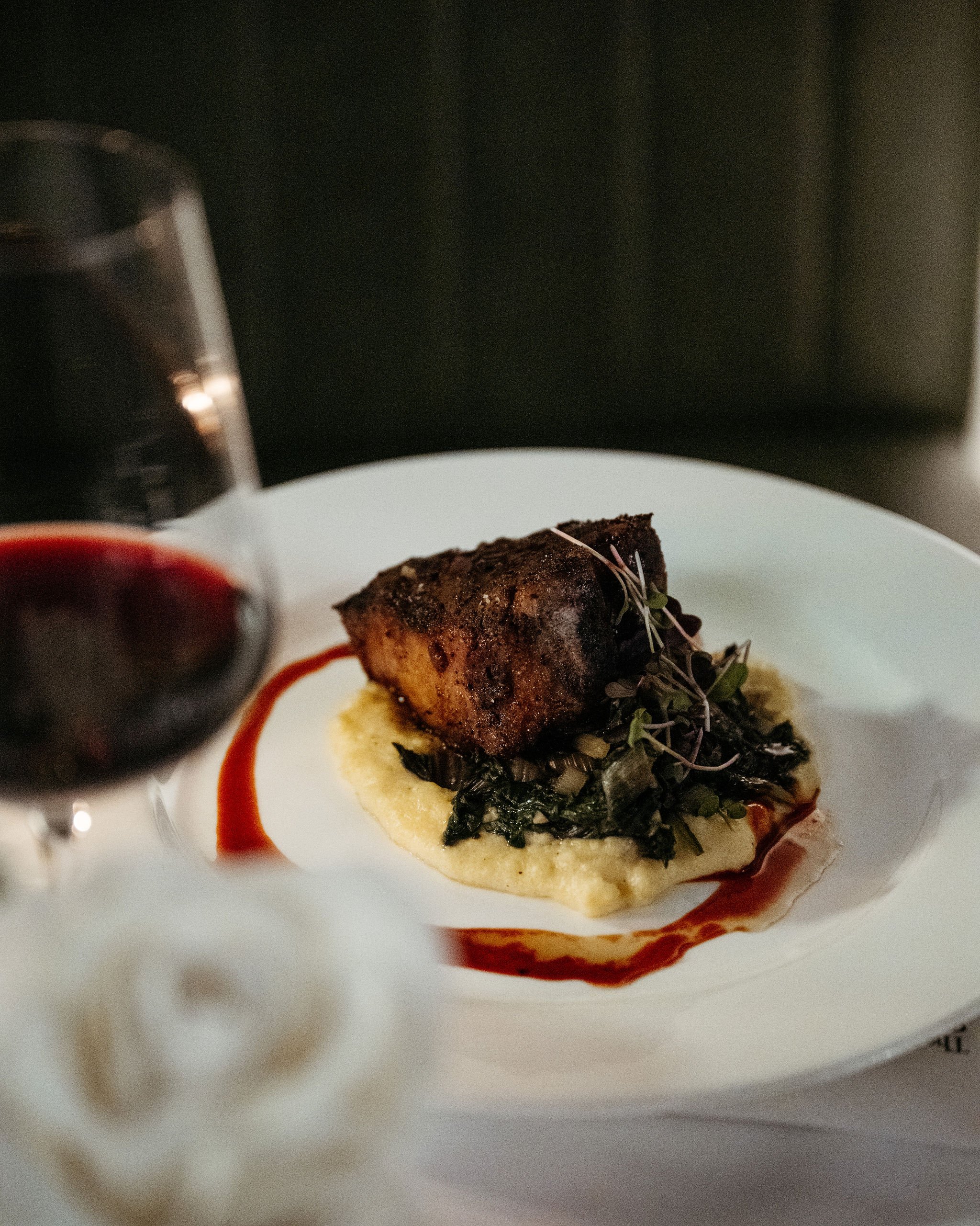 A plated gourmet dish featuring a seared piece of fish on a bed of sautéed greens and mashed potatoes, garnished with microgreens, with a glass of red wine and a dollop of creates in the foreground.