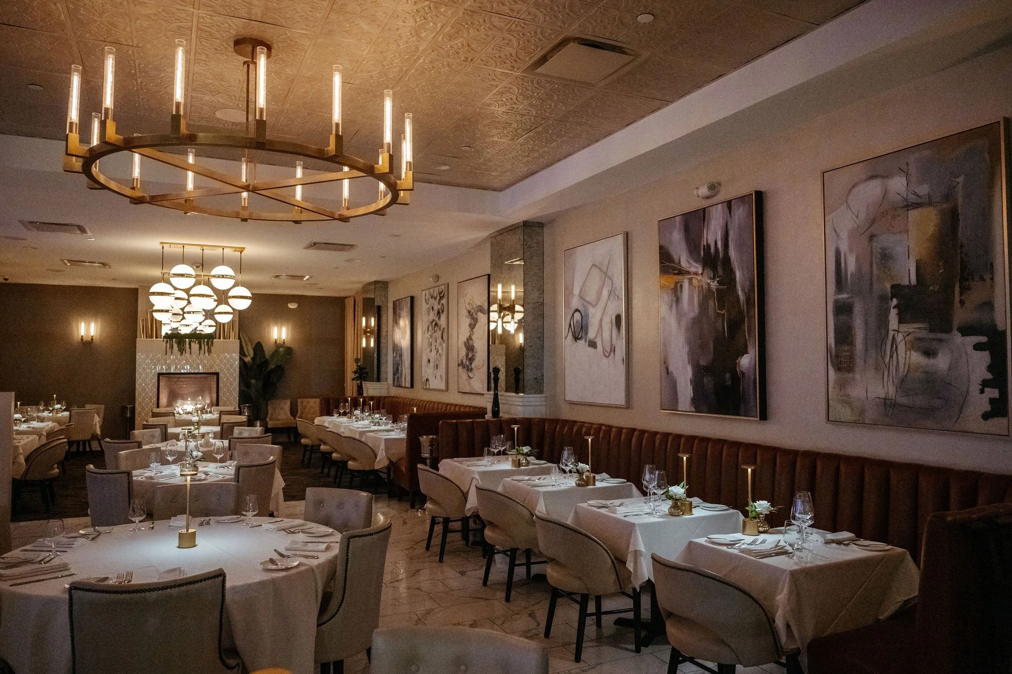 Elegant dining area with multiple round and rectangular tables covered with white tablecloths, set with glassware, silverware, and small floral centerpieces, adorned with modern chandeliers and artwork on the walls.