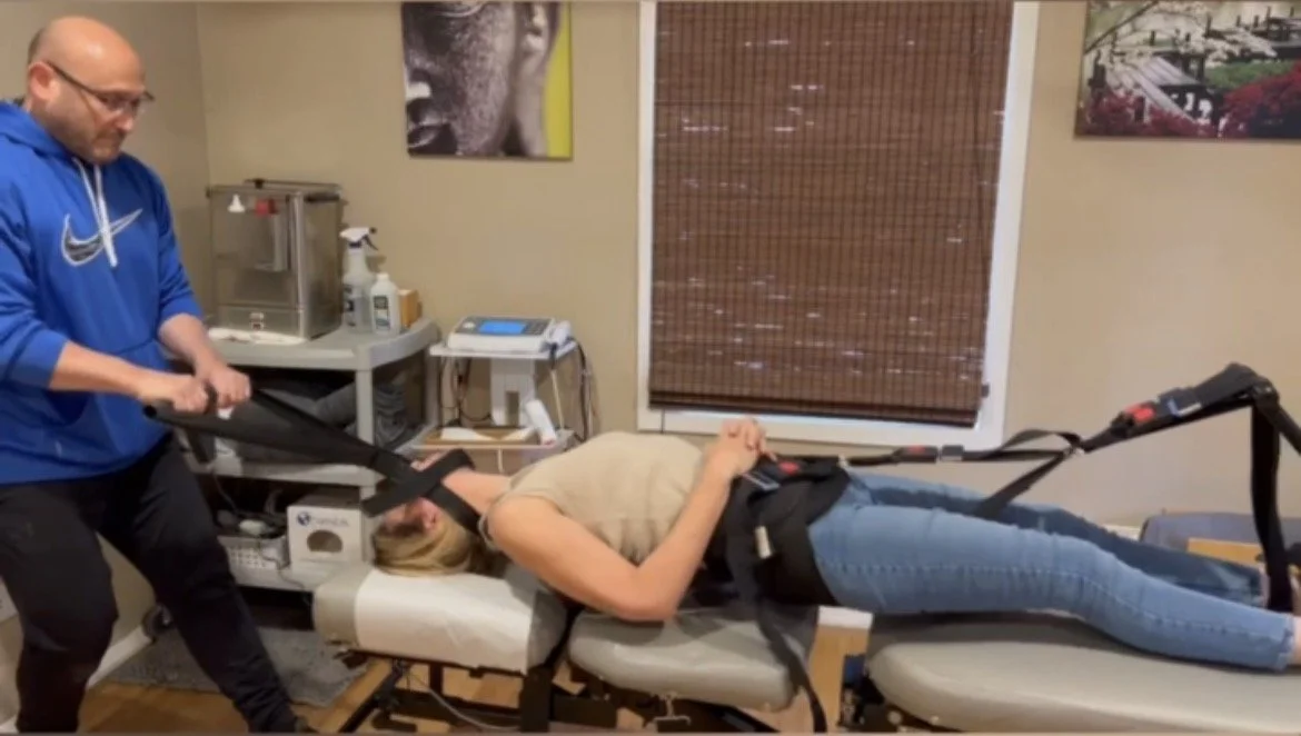 A woman lying on a medical examination table with her hands resting on her stomach, secured with straps, while a man in a blue hoodie adjusts the straps. The room has medical equipment and decor on the walls.