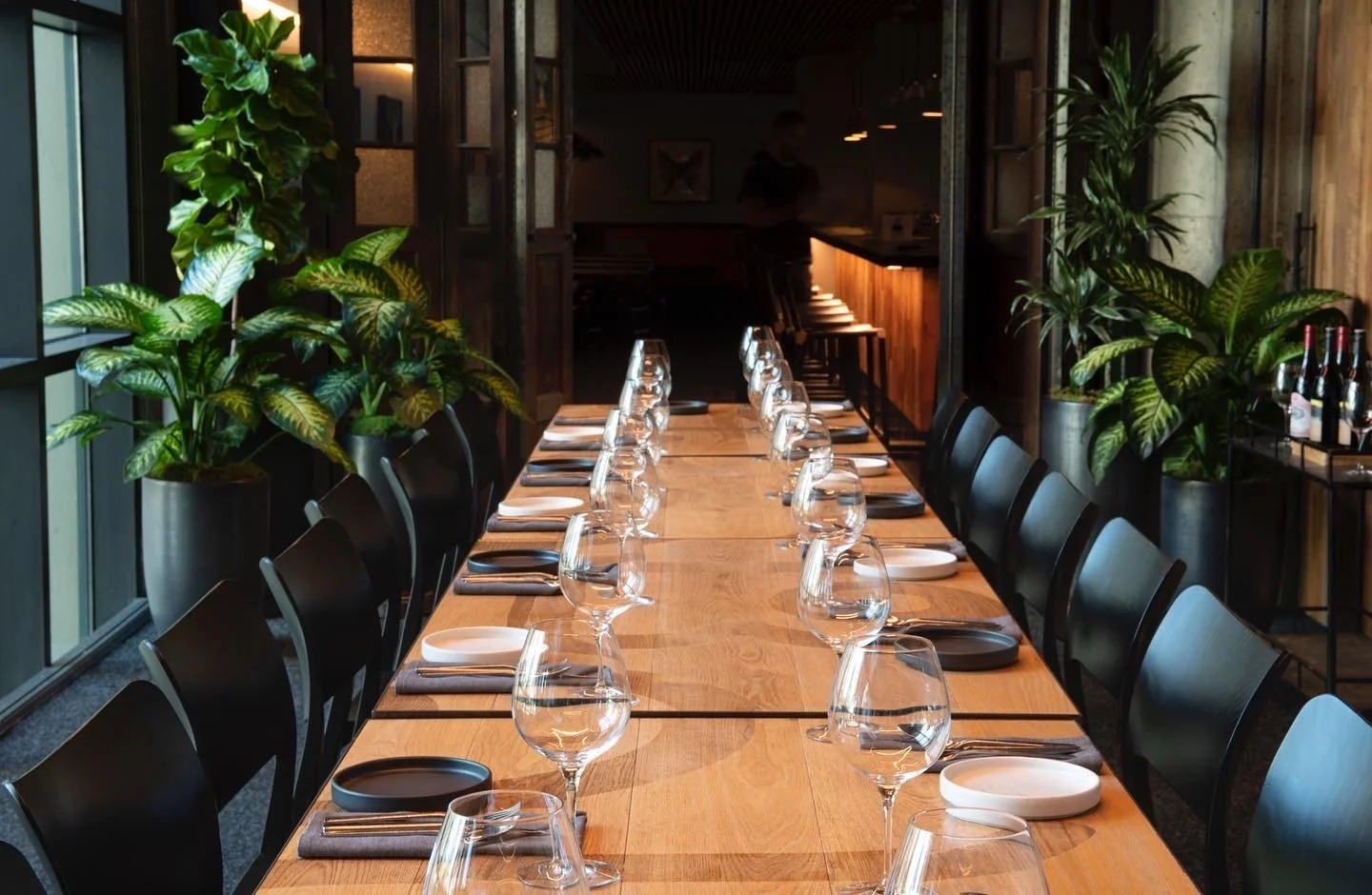 a long wooden table extending forward, flanked by large tropical looking plants; table is set with wine glasses and modern styled dinnerware.