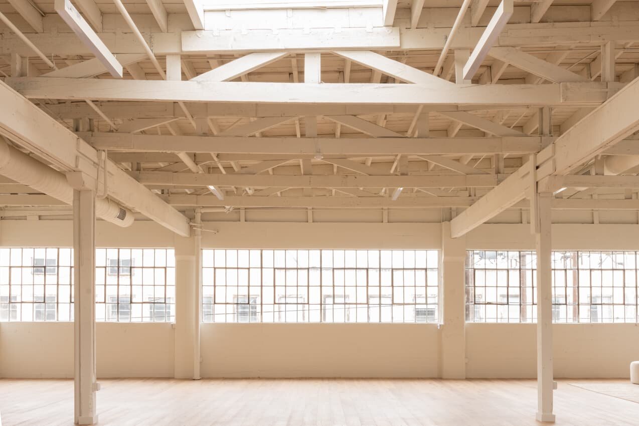 Bright, spacious room with large row of windows, white-painted wooden ceiling beams, and light wood flooring.
