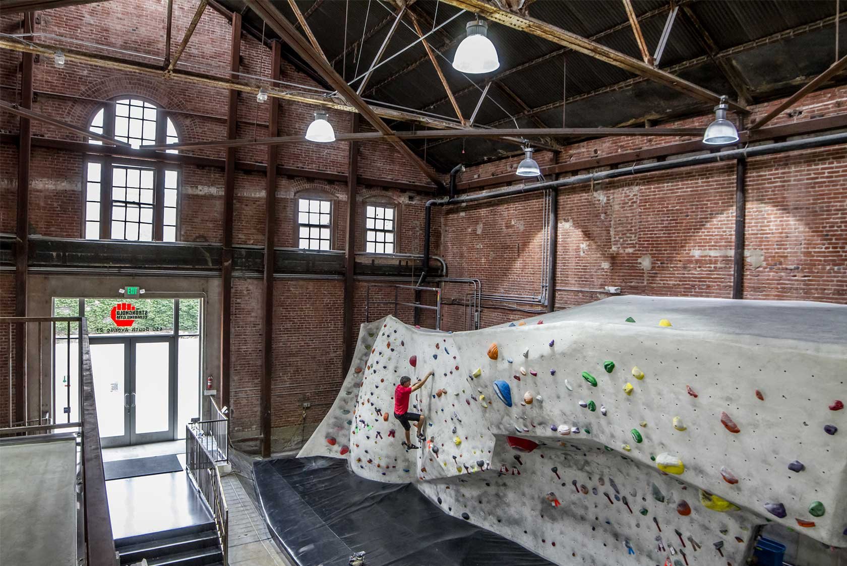 Indoor rock climbing gym with a climber wearing a red shirt climbing a large white wall with colorful holds. The gym features exposed brick walls, large windows, and industrial-style lighting.