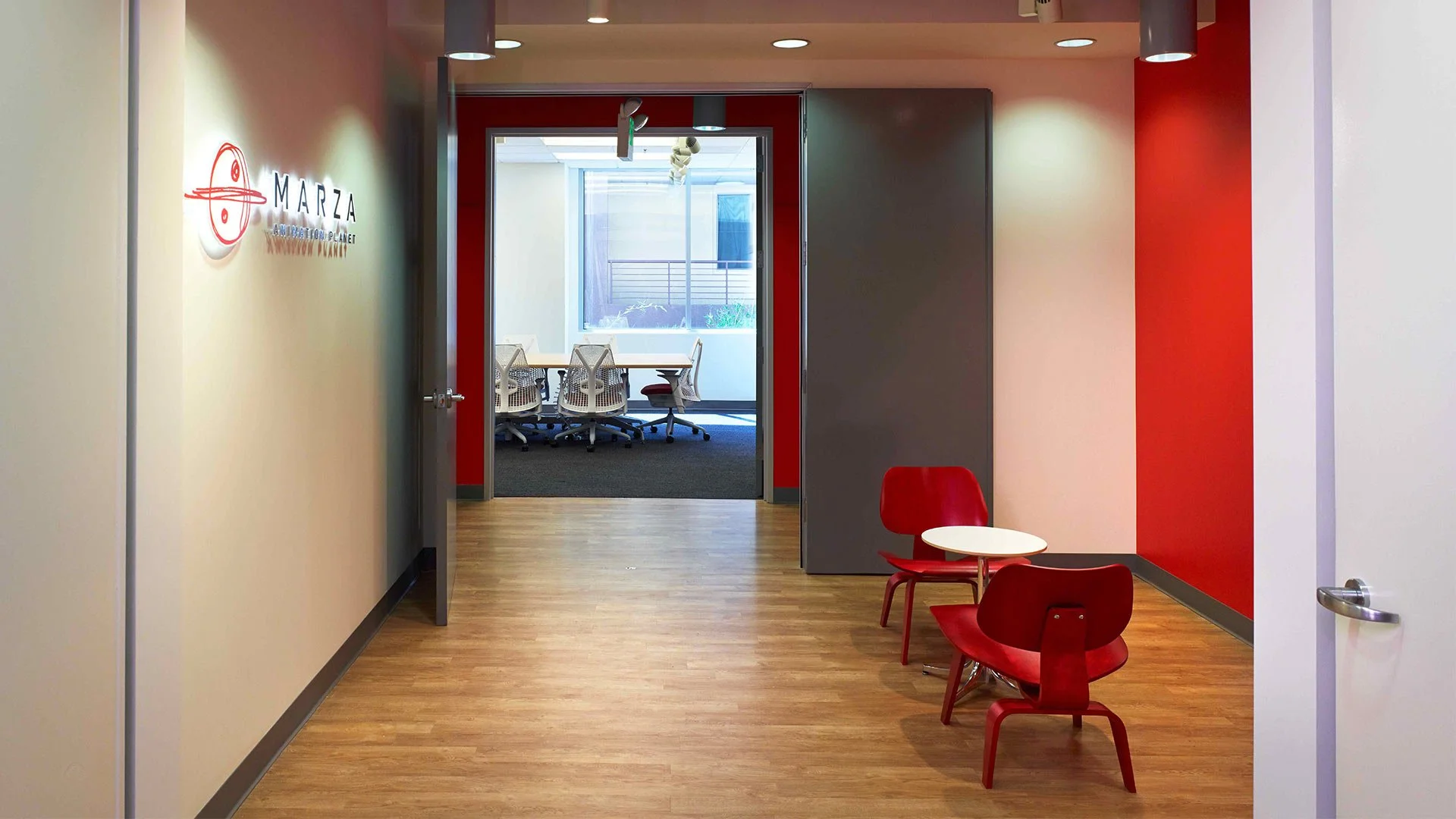 Office hallway leading to a conference room with chairs and a table, featuring red, gray, and white walls, and a wooden floor.