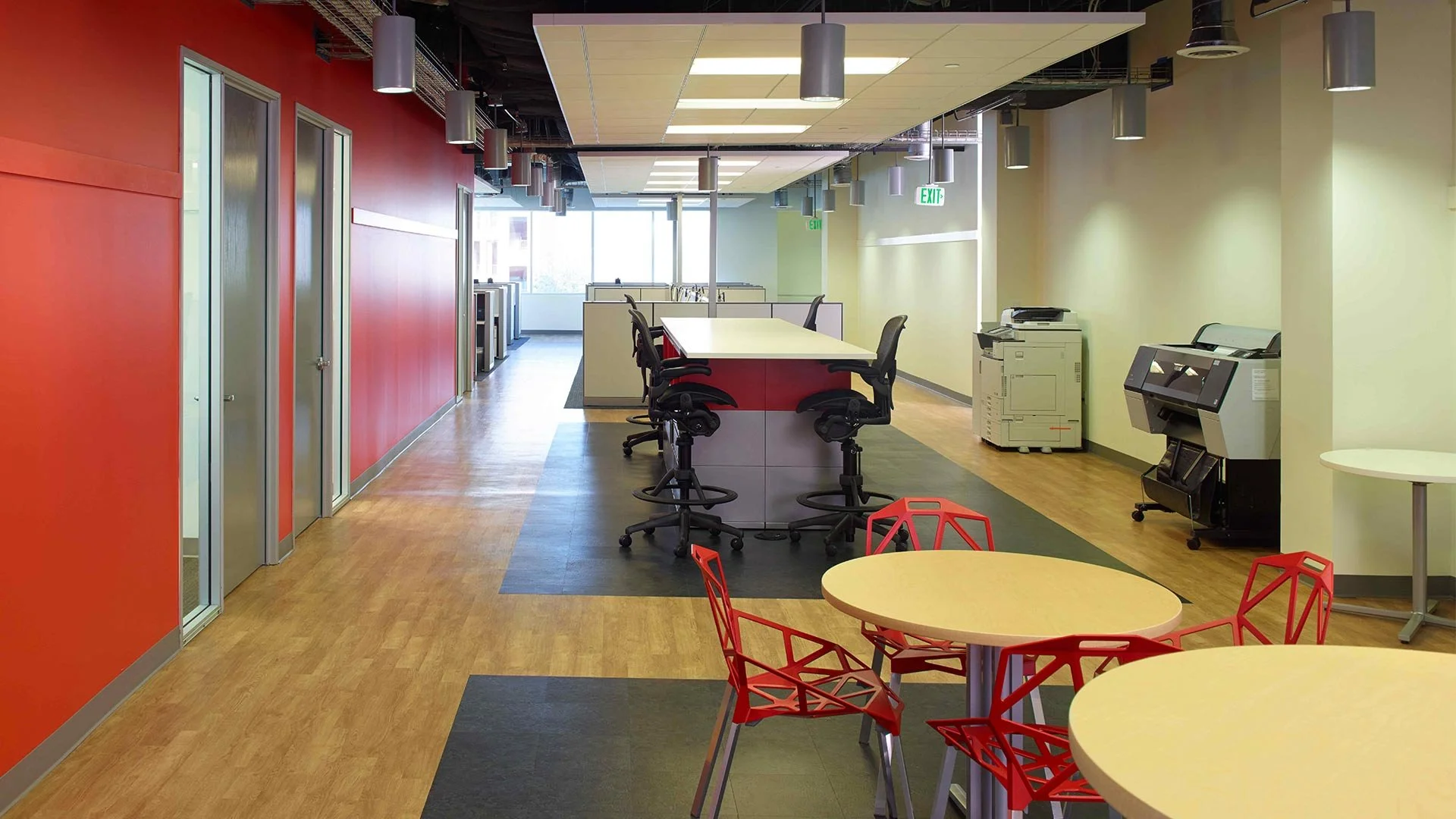 Modern office break room with red, yellow, and green walls, tables, chairs, and office equipment including a copier and printer. Large windows let in natural light.