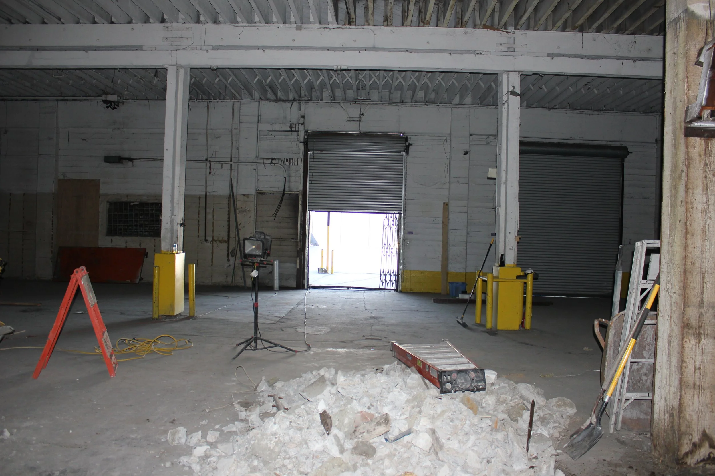 Interior of an unfinished industrial warehouse with exposed beams, partially open garage doors, construction tools, and debris on the floor.