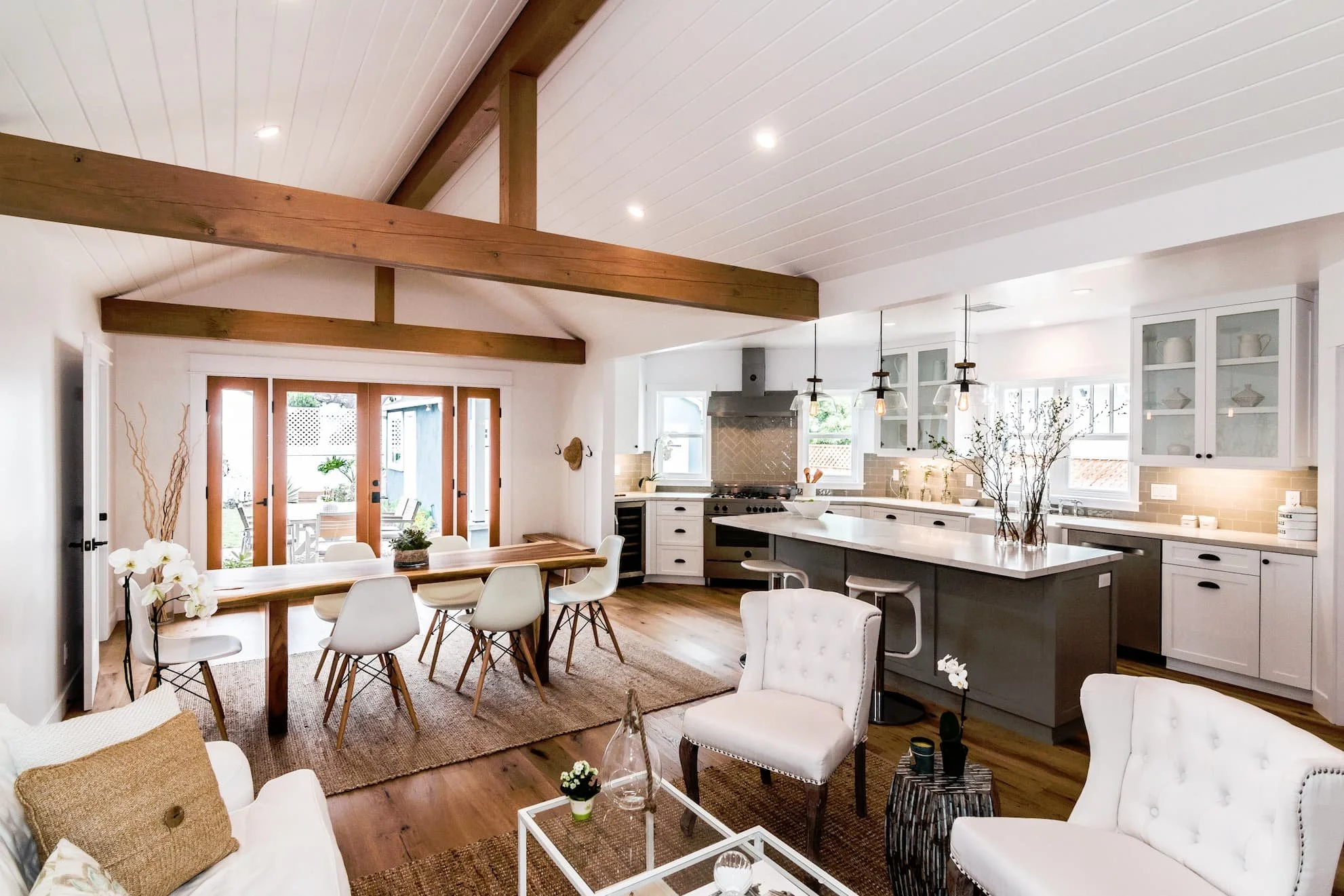 Open-concept living room and kitchen with white walls, wooden beams, hardwood floor, dining table with white chairs, white and gray kitchen cabinets, island with bar stools, and large windows.
