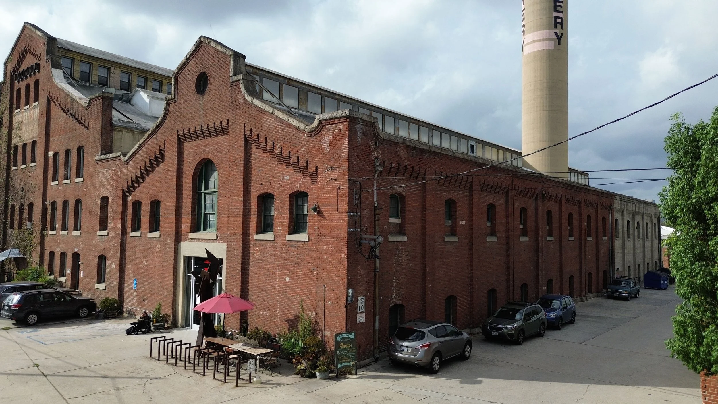 A vintage brick building with arched windows and a tall chimney in the background, with parked cars and a small patio area with umbrellas in the foreground.