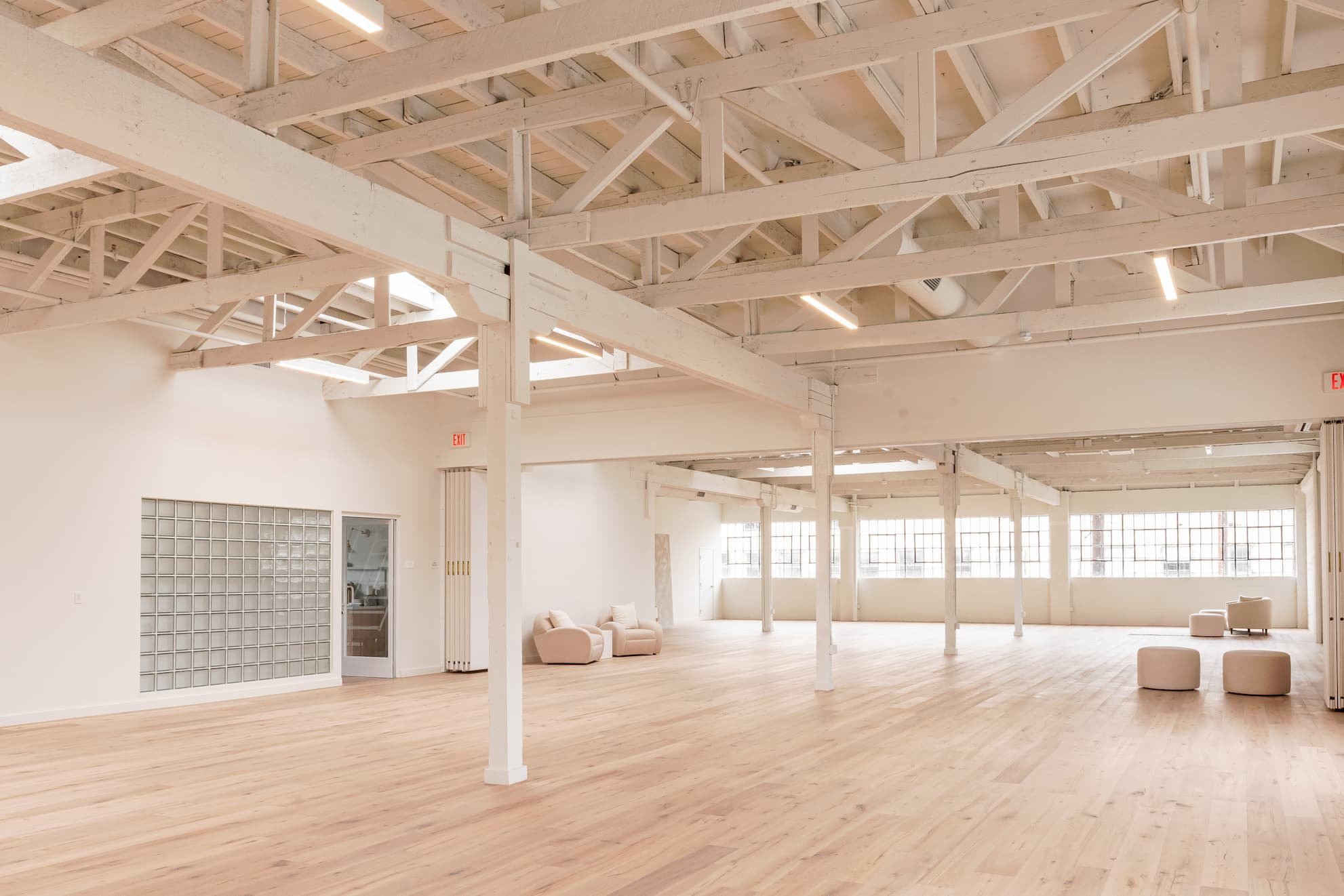 Empty spacious room with high ceilings, exposed white wooden beams, large windows, light wooden flooring, and white walls, with a few beige chairs and ottomans.