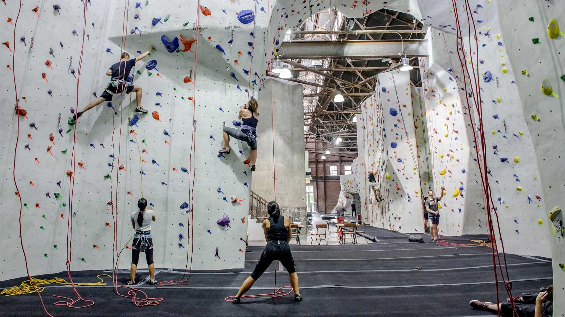 Indoor rock climbing gym with climbers ascending colorful artificial walls, spotting and belaying, and various climbing holds and ropes visible.