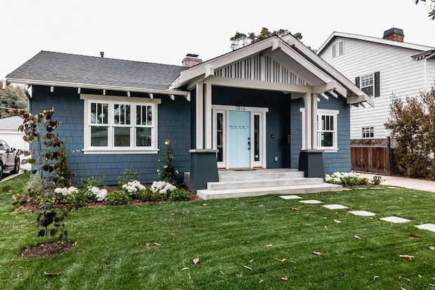 Front view of a blue Craftsman-style house with white trim, a covered porch, and a well-maintained lawn with stepping stones.