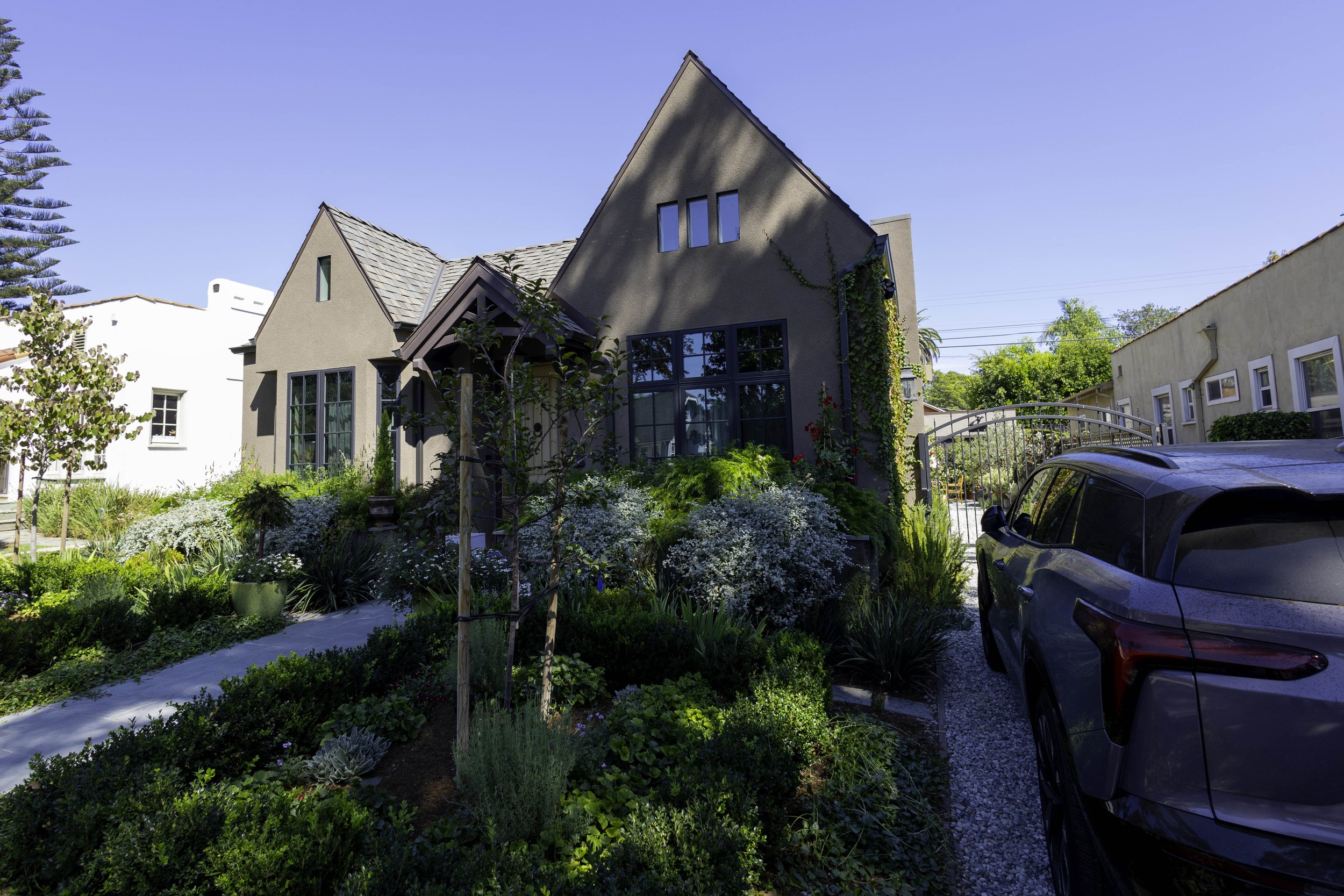 A modern house with large windows and sloped roofs, surrounded by a well-maintained garden with various plants and flowers. A car is parked in the driveway, and the sky is clear and blue.