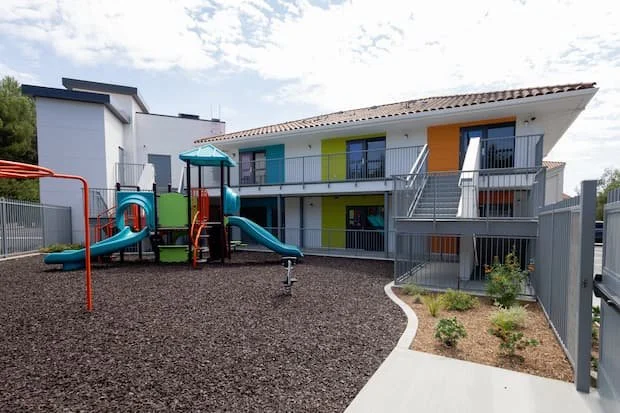Colorful playground with slides and a shaded area, surrounded by a modern apartment building with colorful doors and balconies.