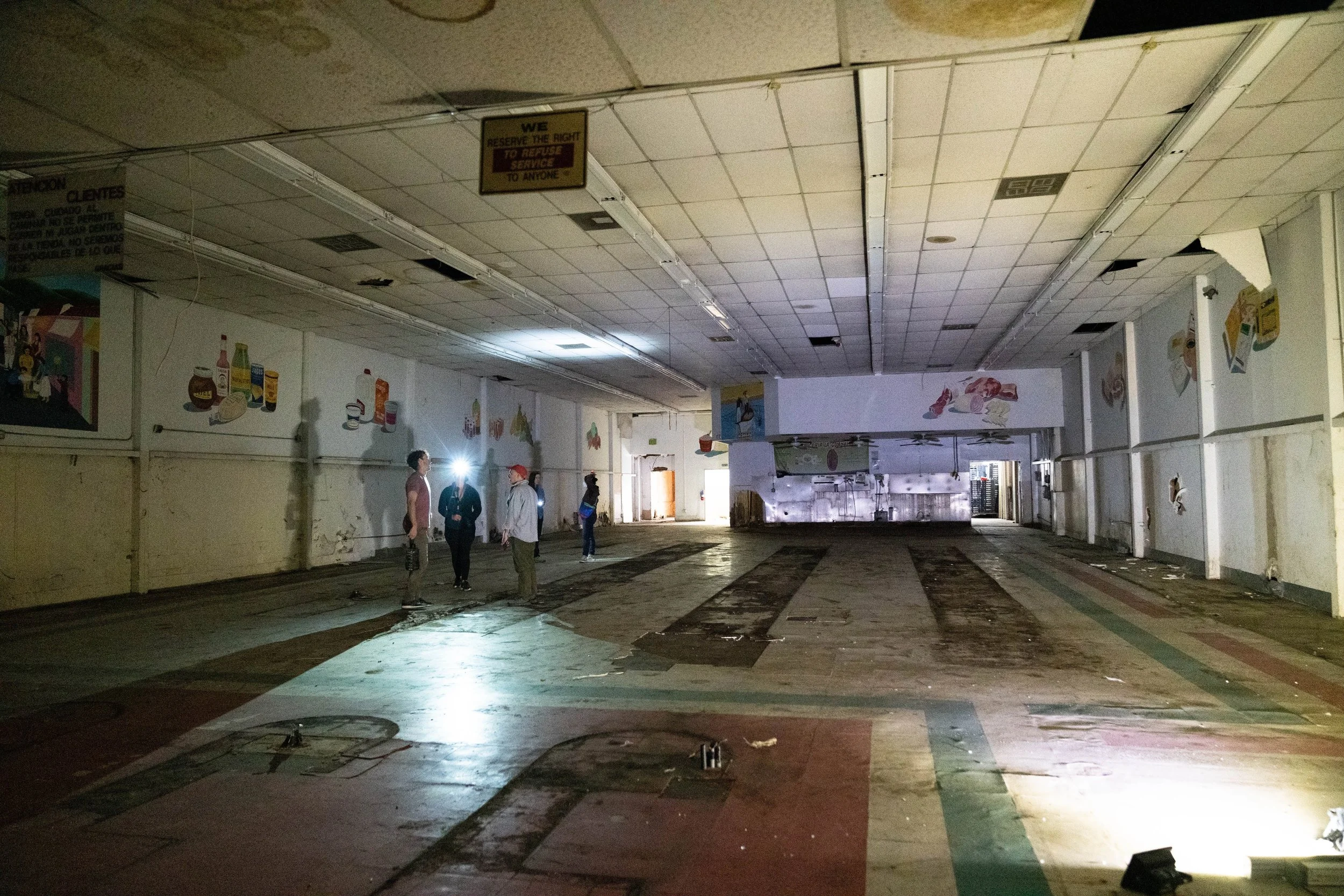An interior of an abandoned store or shopping mall with a damaged ceiling, a few people standing and talking, some lights on, broken tiles on the floor, and promotional posters on the walls.