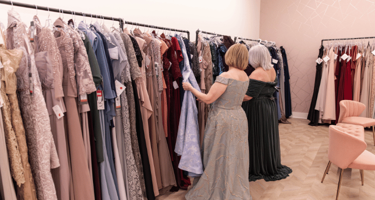 Two women in elegant gowns browse at Heart to Heart Bride's mother of the groom dress selection. Racks are filled with assorted styles and colors, exuding a sense of luxury.