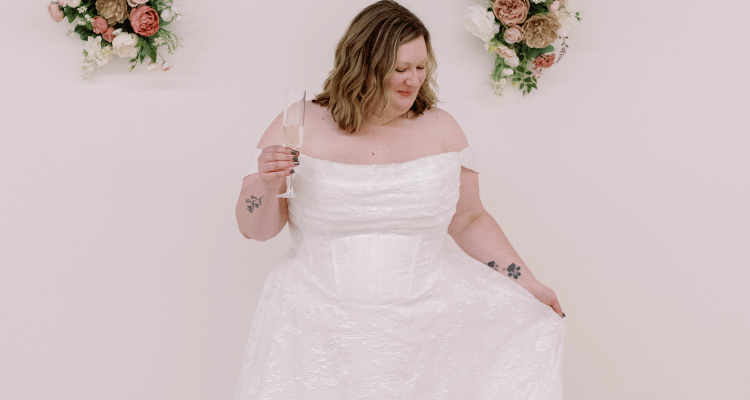 Bride holding a champagne glass and smiling in an off-the-shoulder satin wedding gown with a structured corset bodice and full skirt.